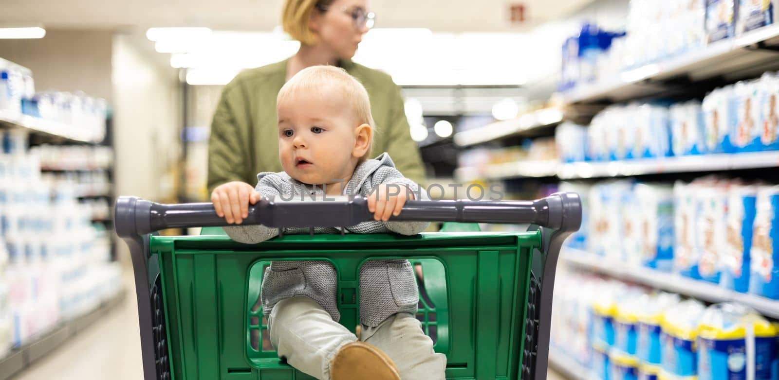 Mother pushing shopping cart with her infant baby boy child down department aisle in supermarket grocery store. Shopping with kids concept. by kasto