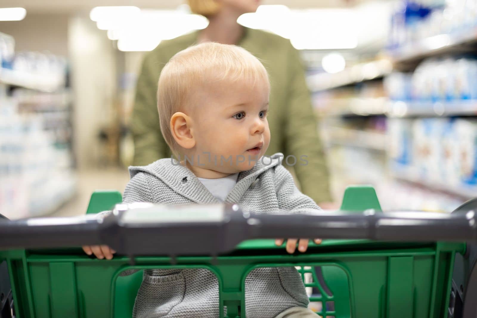 Mother pushing shopping cart with her infant baby boy child down department aisle in supermarket grocery store. Shopping with kids concept
