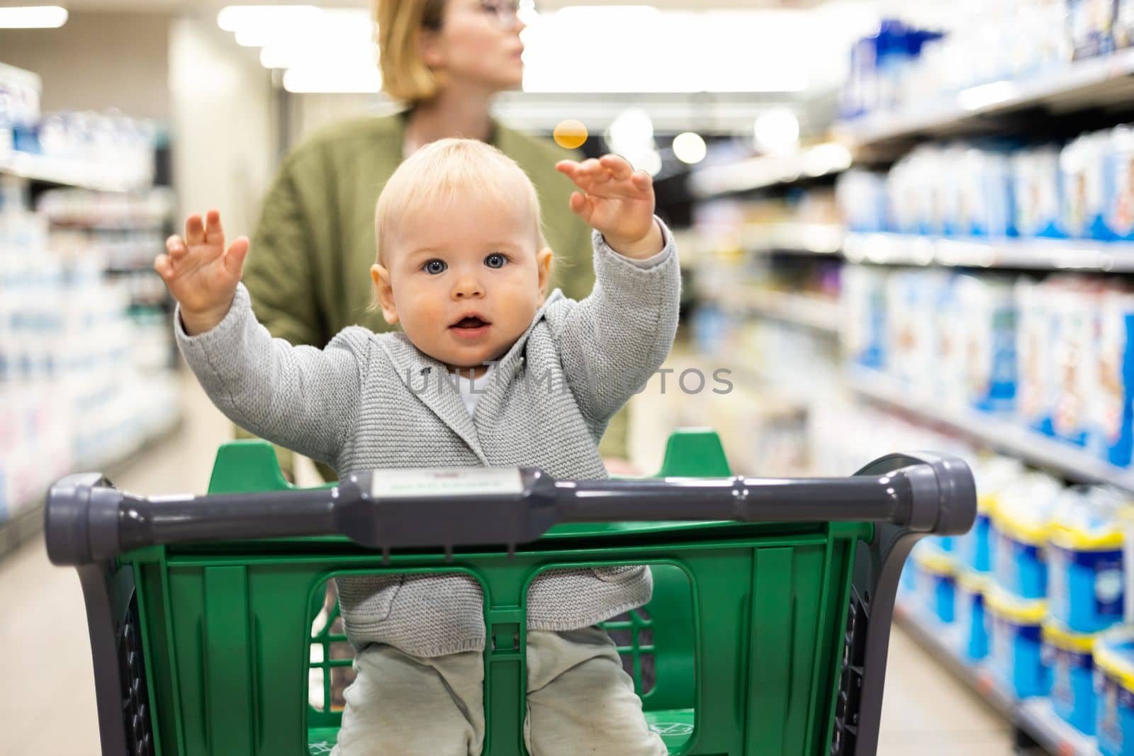 Mother pushing shopping cart with her infant baby boy child down department aisle in supermarket grocery store. Shopping with kids concept. by kasto