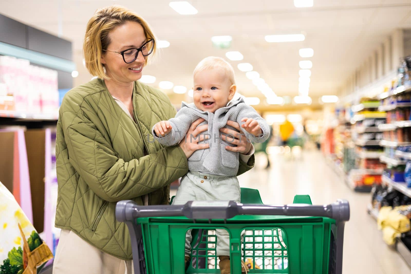 Mother pushing shopping cart with her infant baby boy child down department aisle in supermarket grocery store. Shopping with kids concept