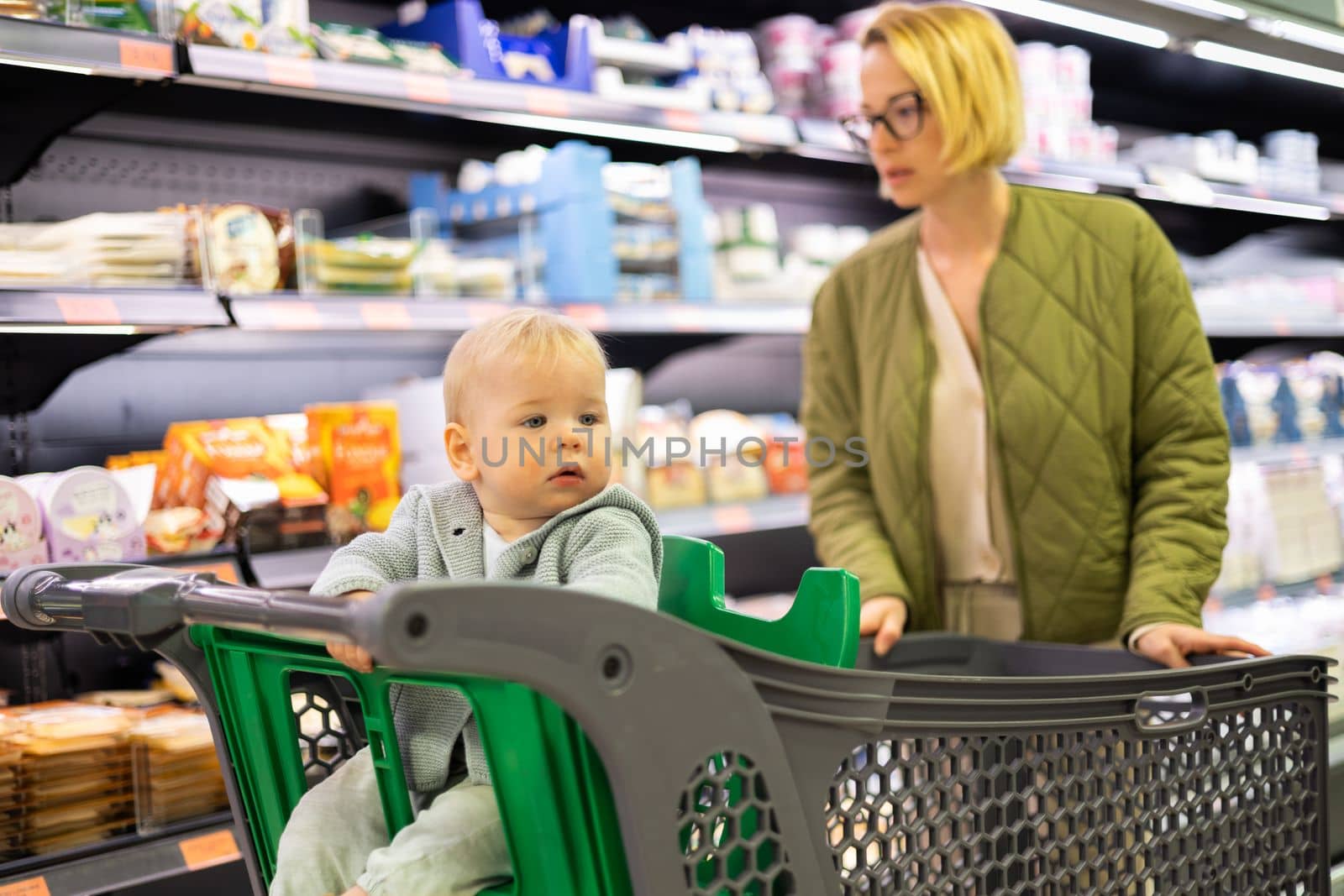 Casualy dressed mother choosing canned products in department of supermarket grocery store with her infant baby boy child in shoping cart by kasto