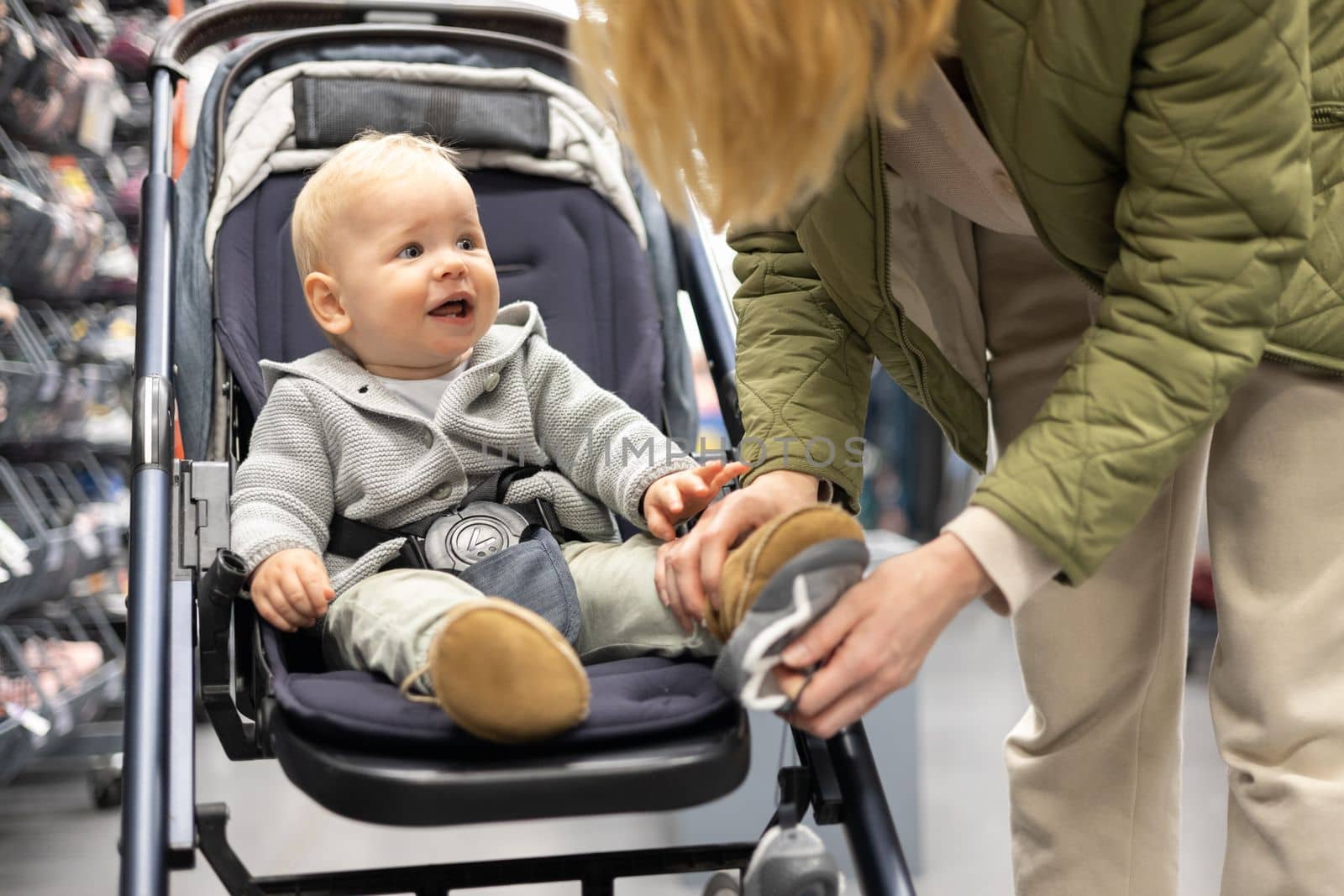 Casualy dressed mother choosing sporty shoes and clothes products in sports department of supermarket store with her infant baby boy child in stroller