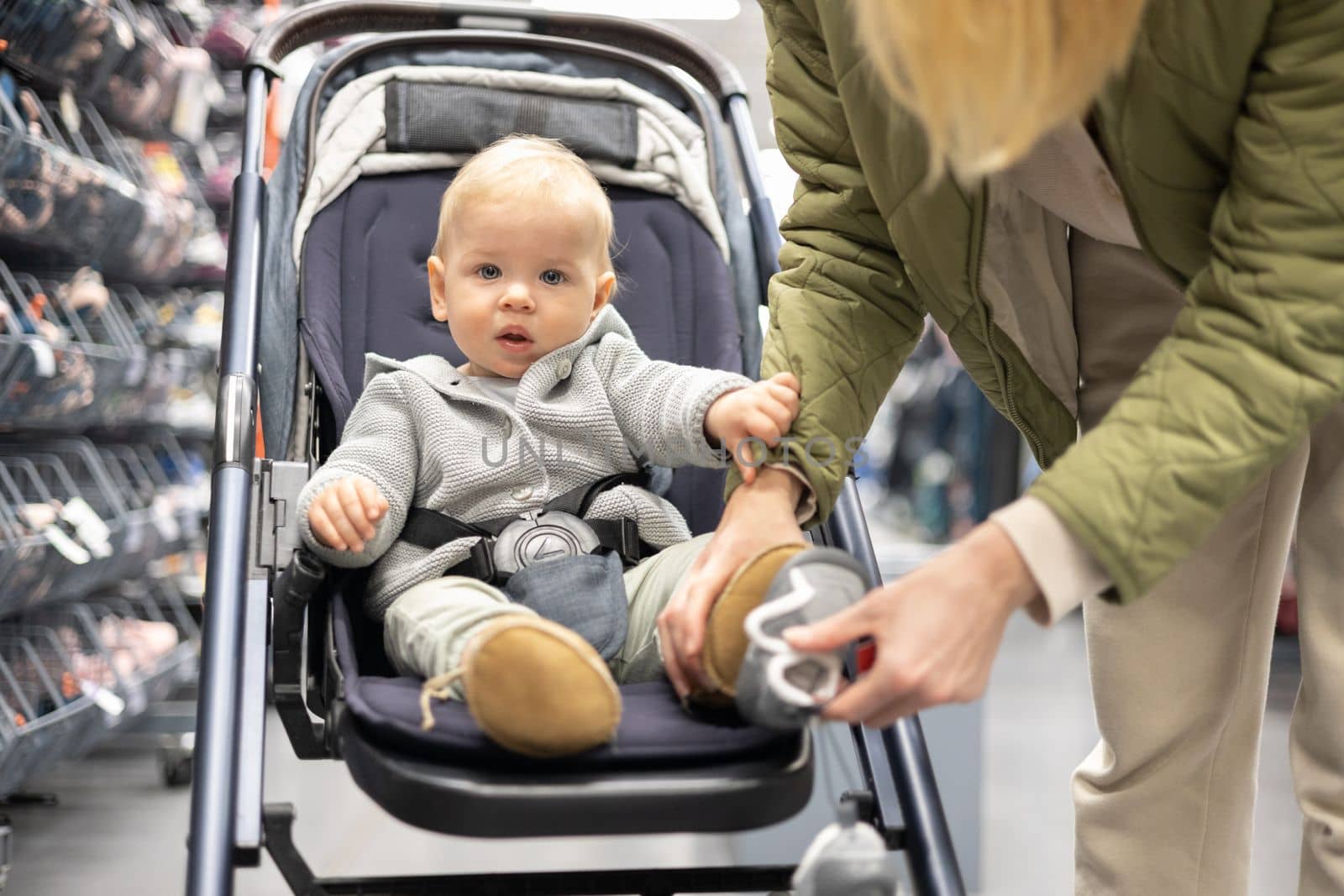 Casualy dressed mother choosing sporty shoes and clothes products in sports department of supermarket store with her infant baby boy child in stroller. by kasto