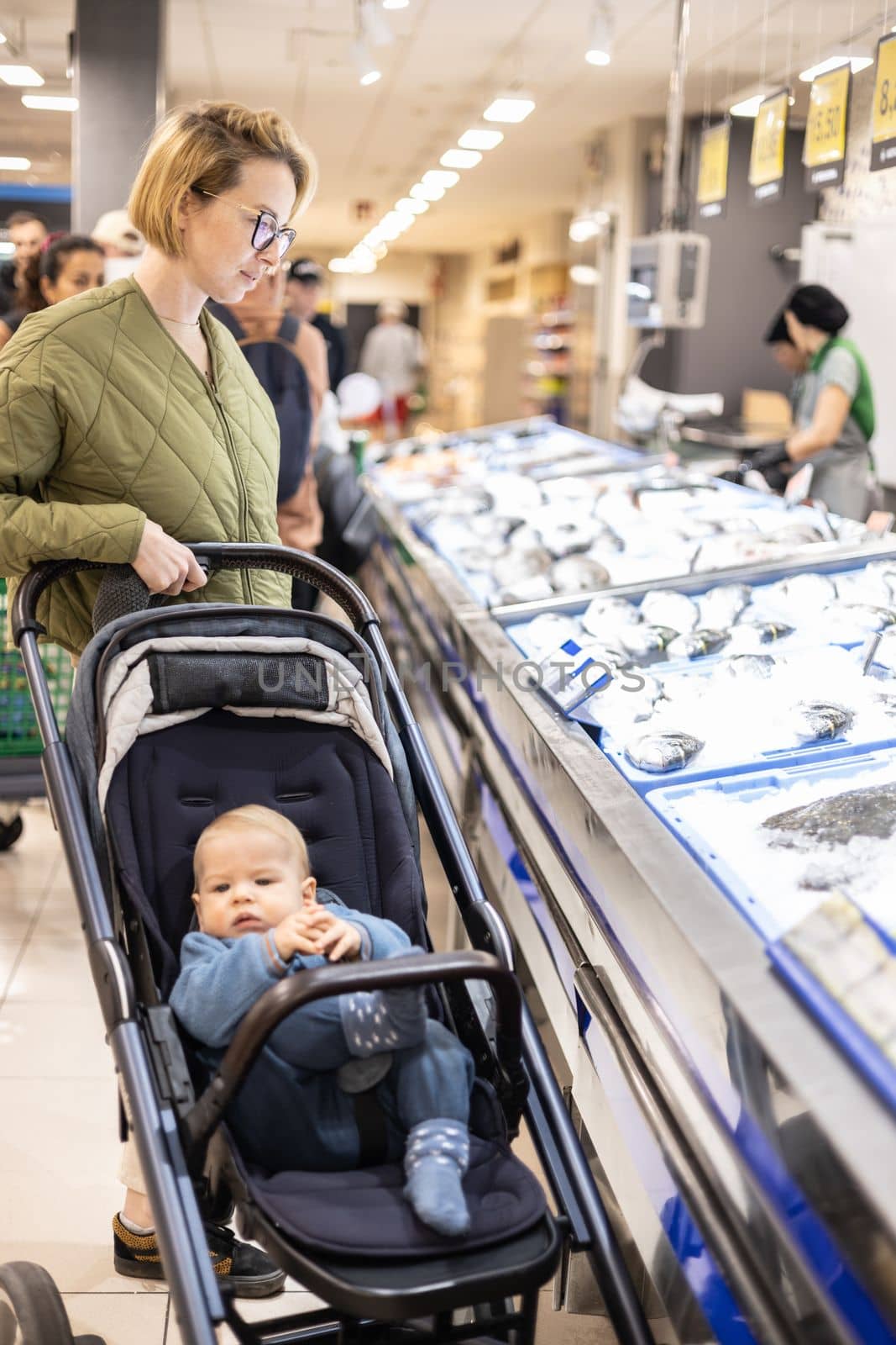 Casualy dressed mother choosing fish in the fish market department of supermarket grocery store with her infant baby boy child in stroller