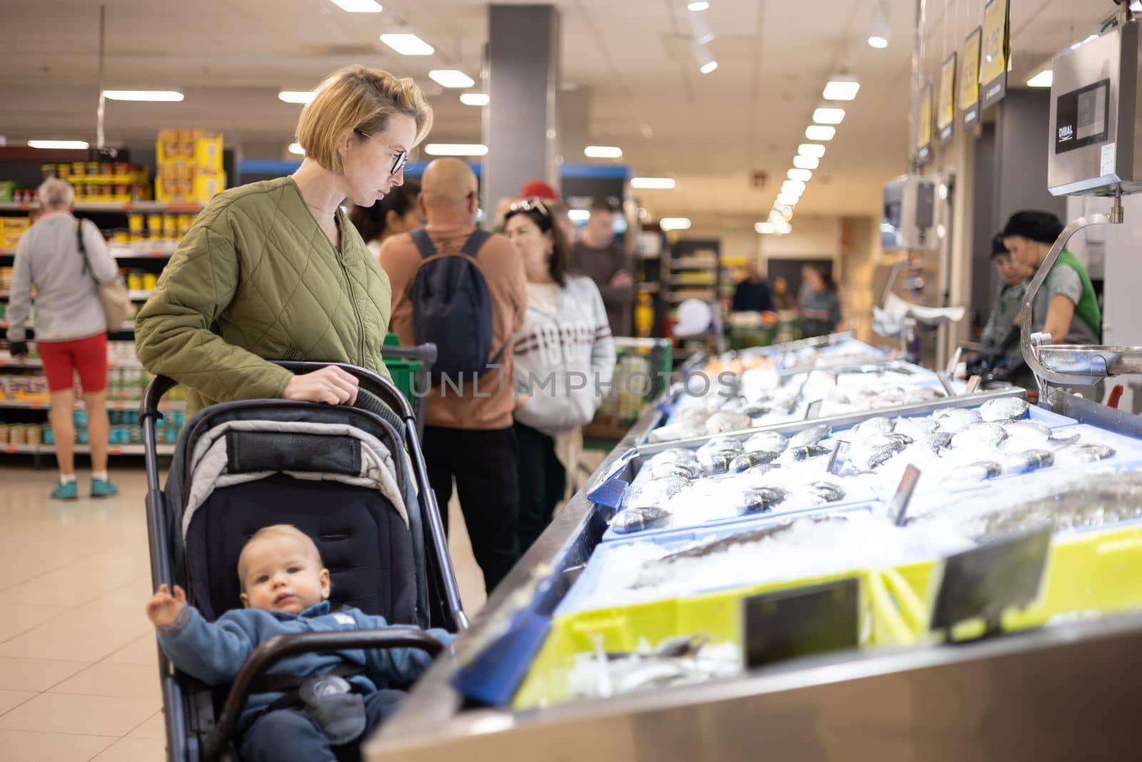 Casualy dressed mother choosing fish in the fish market department of supermarket grocery store with her infant baby boy child in stroller
