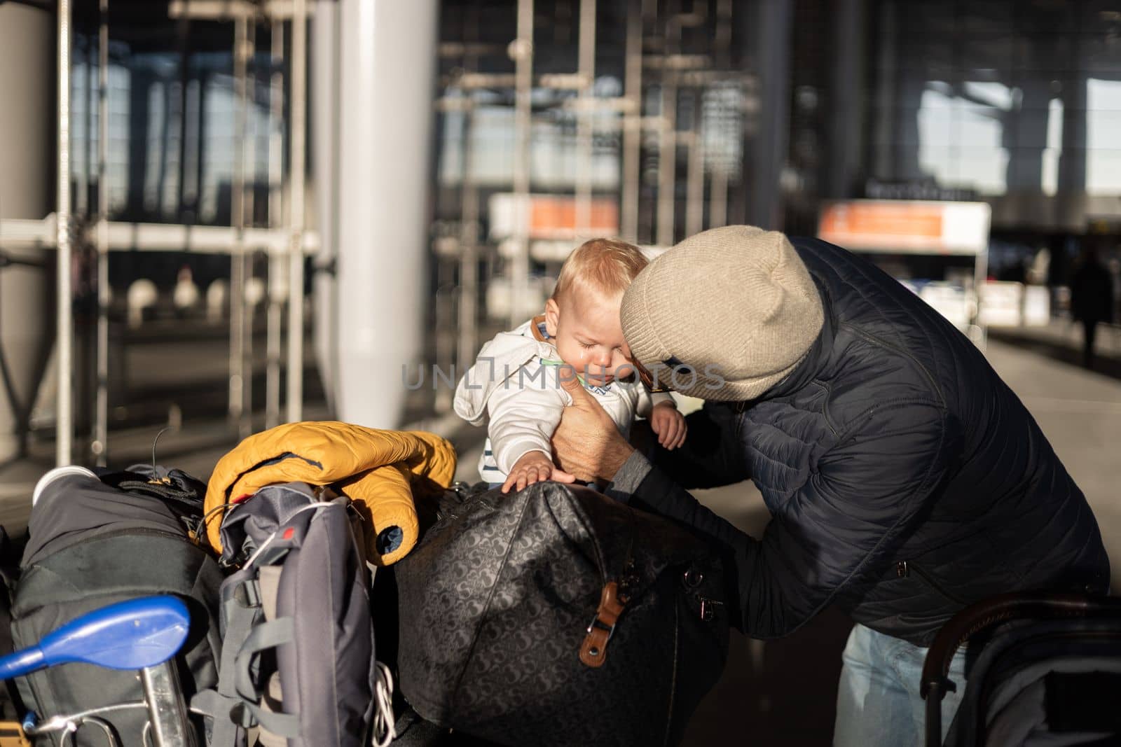 Fatherat comforting his crying infant baby boy child tired sitting on top of luggage cart in front of airport terminal station while traveling wih family