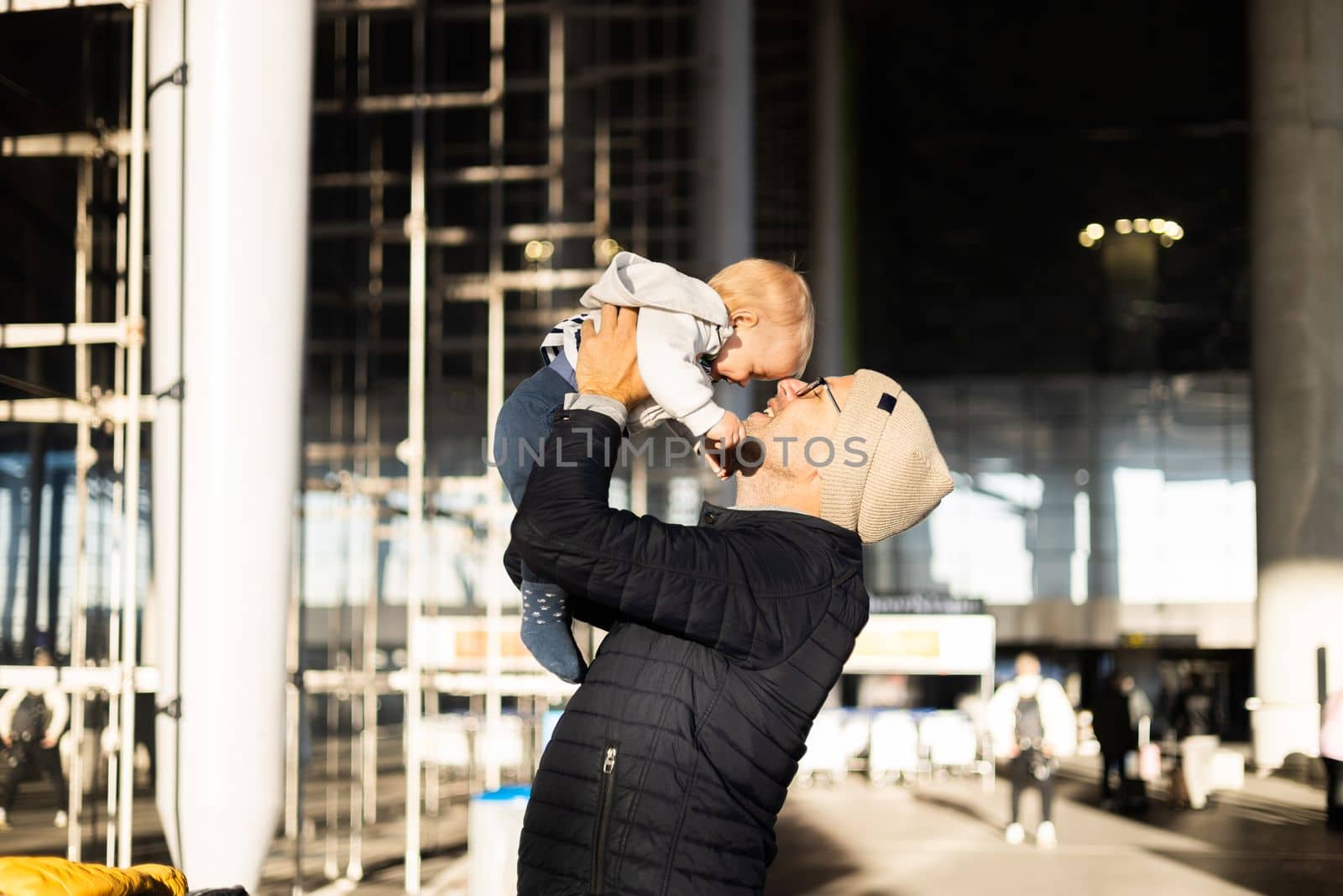 Father happily holding and lifting his infant baby boy child in the air after being rejunited in front of airport terminal station. Baby travel concept