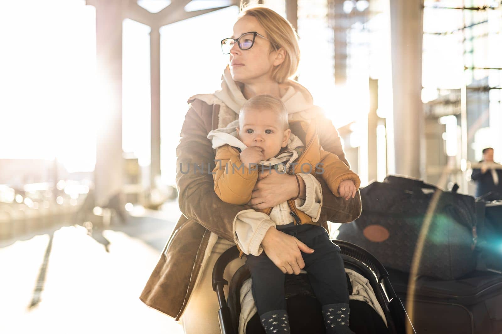 Motherat travelling with his infant baby boy child, walking, pushing baby stroller and luggage cart in front of airport terminal station