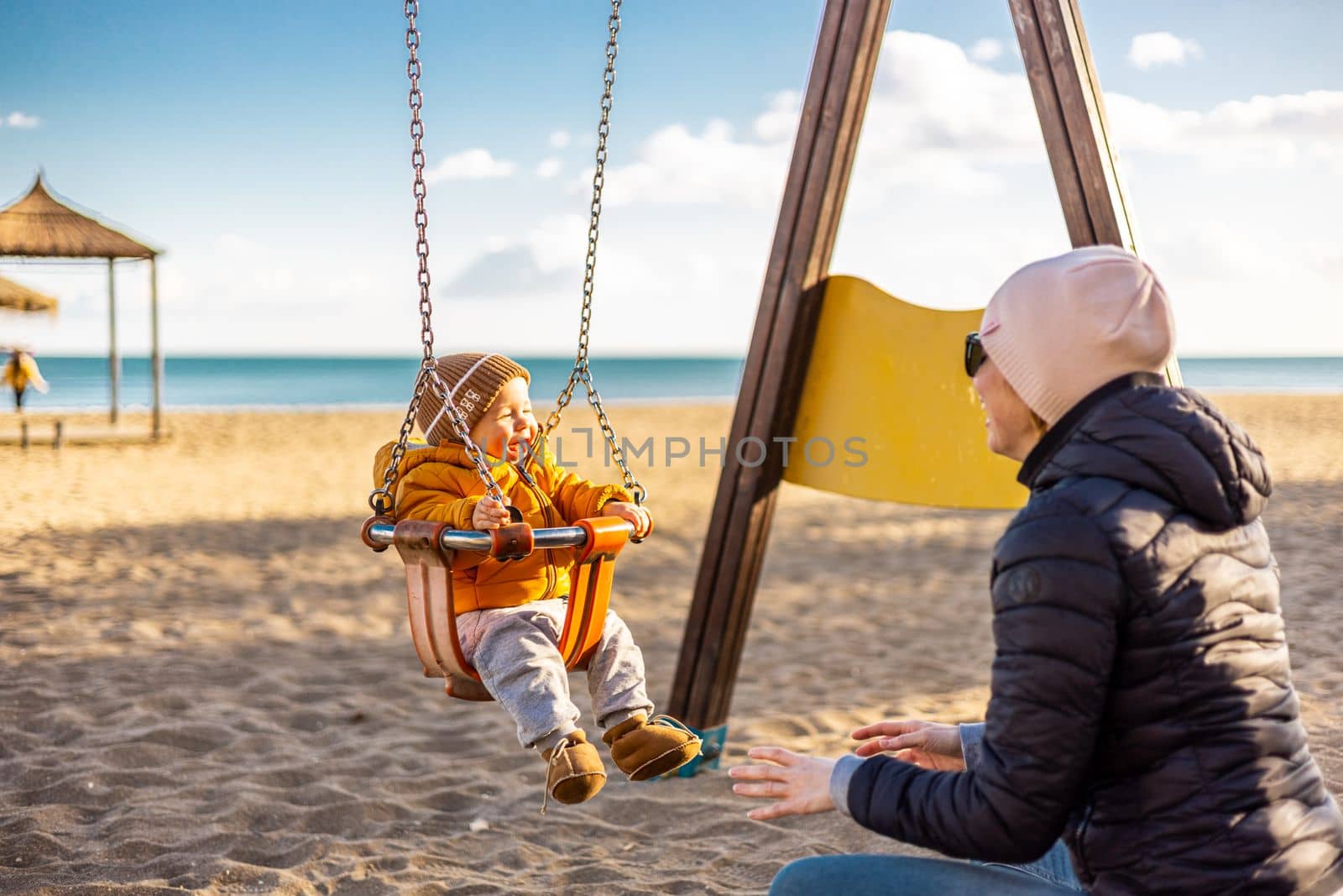 Mother pushing her infant baby boy child on a swing on sandy beach playground outdoors on nice sunny cold winter day in Malaga, Spain. by kasto