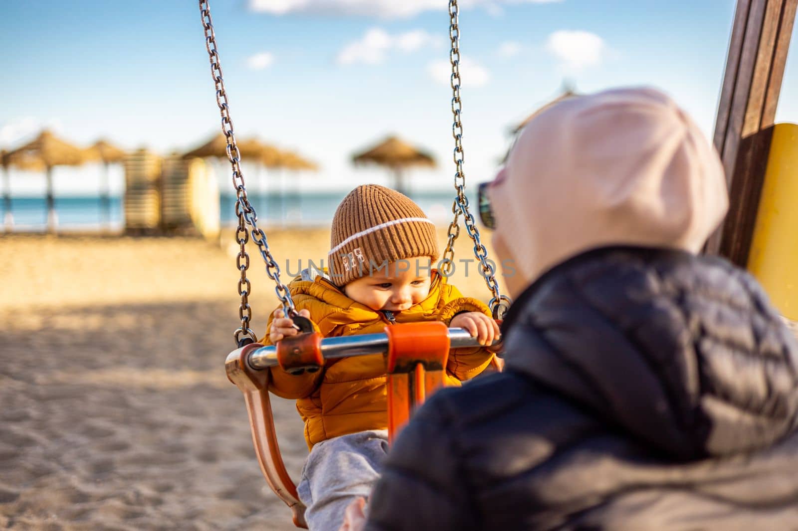 Mother pushing her infant baby boy child on a swing on sandy beach playground outdoors on nice sunny cold winter day in Malaga, Spain. by kasto