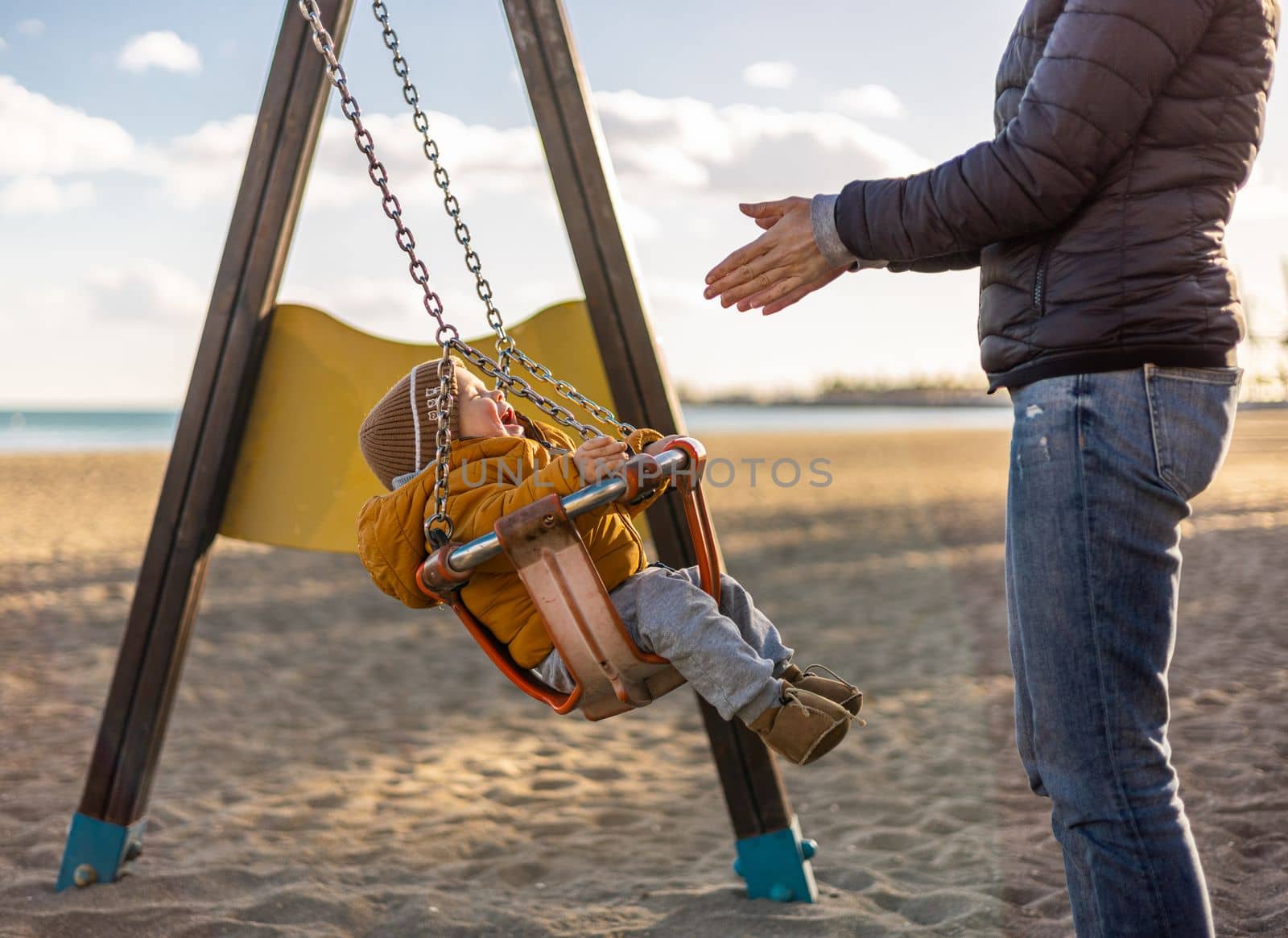 Mother pushing her infant baby boy child on a swing on sandy beach playground outdoors on nice sunny cold winter day in Malaga, Spain. by kasto