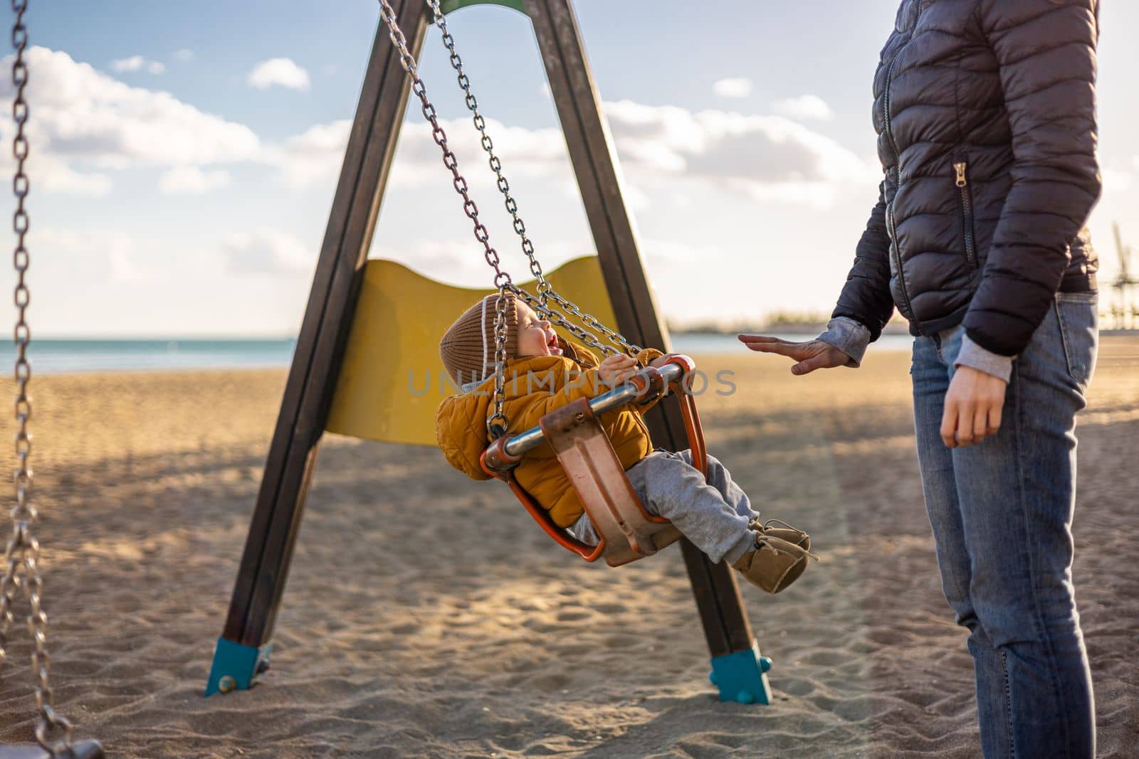 Mother pushing her infant baby boy child on a swing on sandy beach playground outdoors on nice sunny cold winter day in Malaga, Spain. by kasto