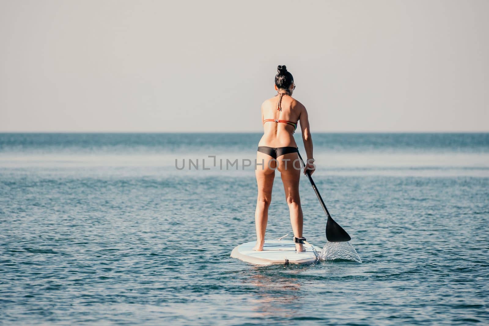 Sea woman sup. Silhouette of happy middle aged tanned woman in rainbow bikini, surfing on SUP board, confident paddling through water surface. Idyllic sunset. Active lifestyle at sea or river. by panophotograph