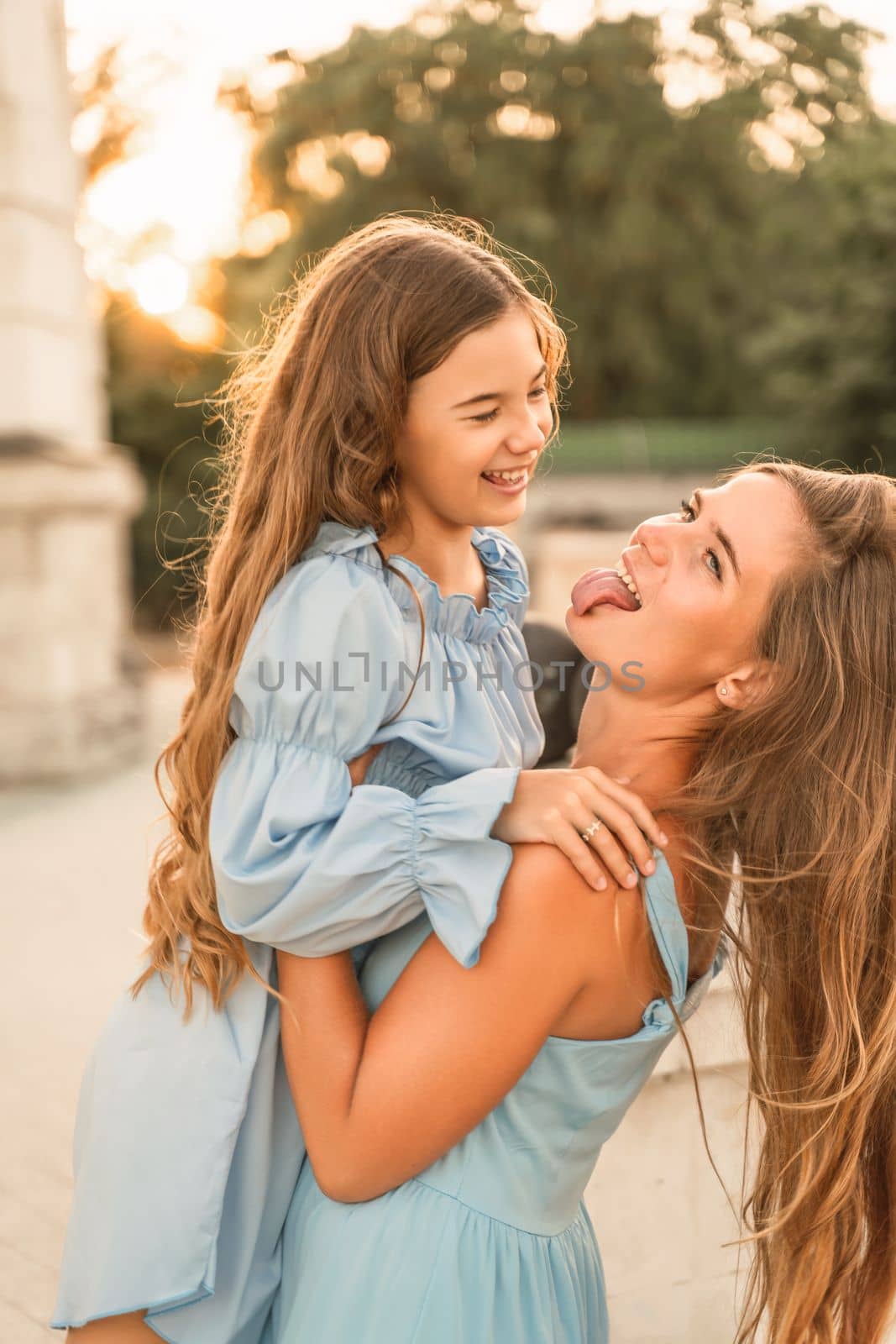 Mother and daughter in blue dresses with flowing long hair against the backdrop of a sunset and a white building. They look at each other. Family stories on the weekend.