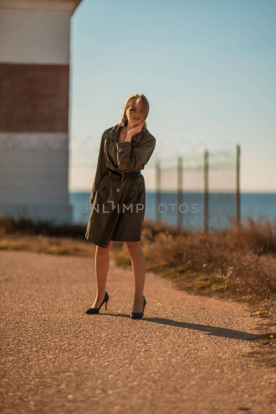 Portrait blonde sea cape. A calm young blonde in a khaki raincoat stands on the seashore against the backdrop of a lighthouse. by Matiunina