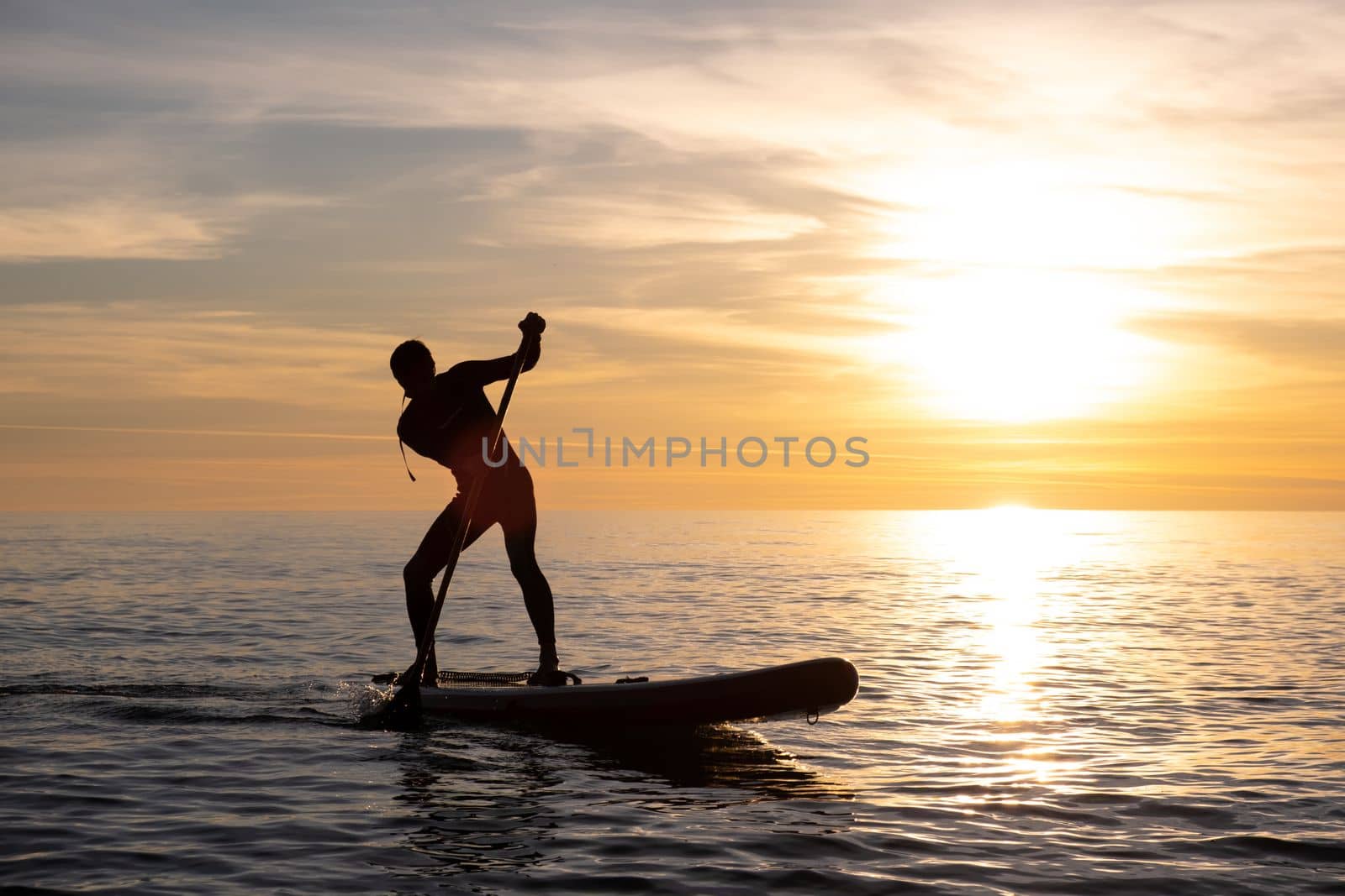 a sporty guy swims with a paddle on a sapboard in the sea under the beautiful sunset sun