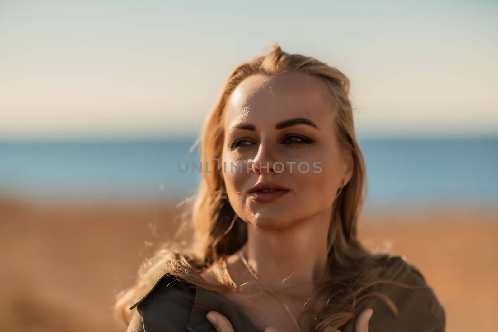 Portrait blonde sea cape. A calm young blonde in an unbuttoned khaki raincoat stands on the seashore, under the raincoat there is a black skirt and top by Matiunina