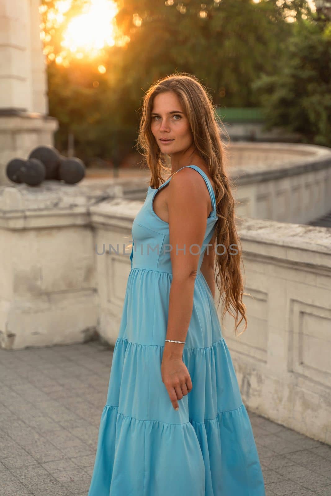 Woman sunset blue dress. Portrait of a woman with long hair and a blue dress against the backdrop of the setting sun and a white building. Lifestyle, walking around the city by Matiunina