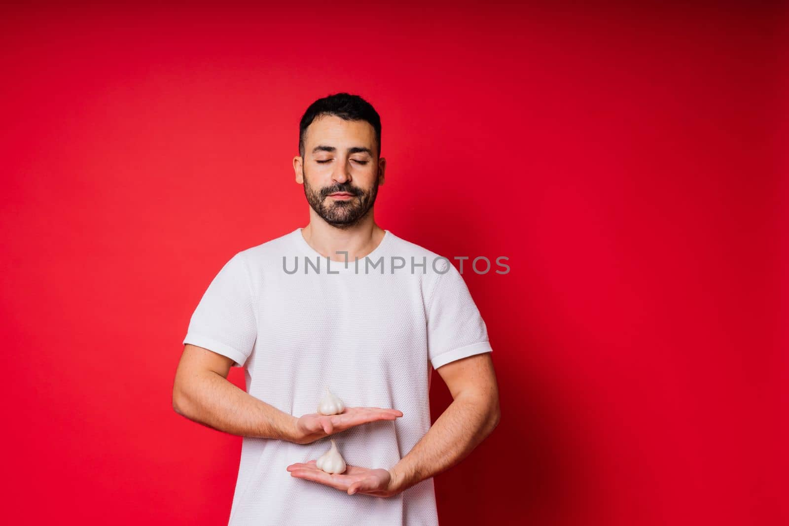 Young bearded man in white shirt is holding garlics with smile, isolated over red background by Zelenin