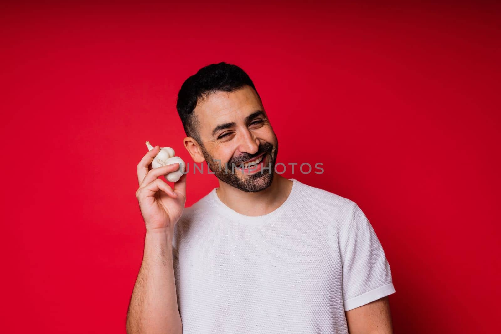 Young bearded man in a white shirt is holding garlics with smile, isolated over red background