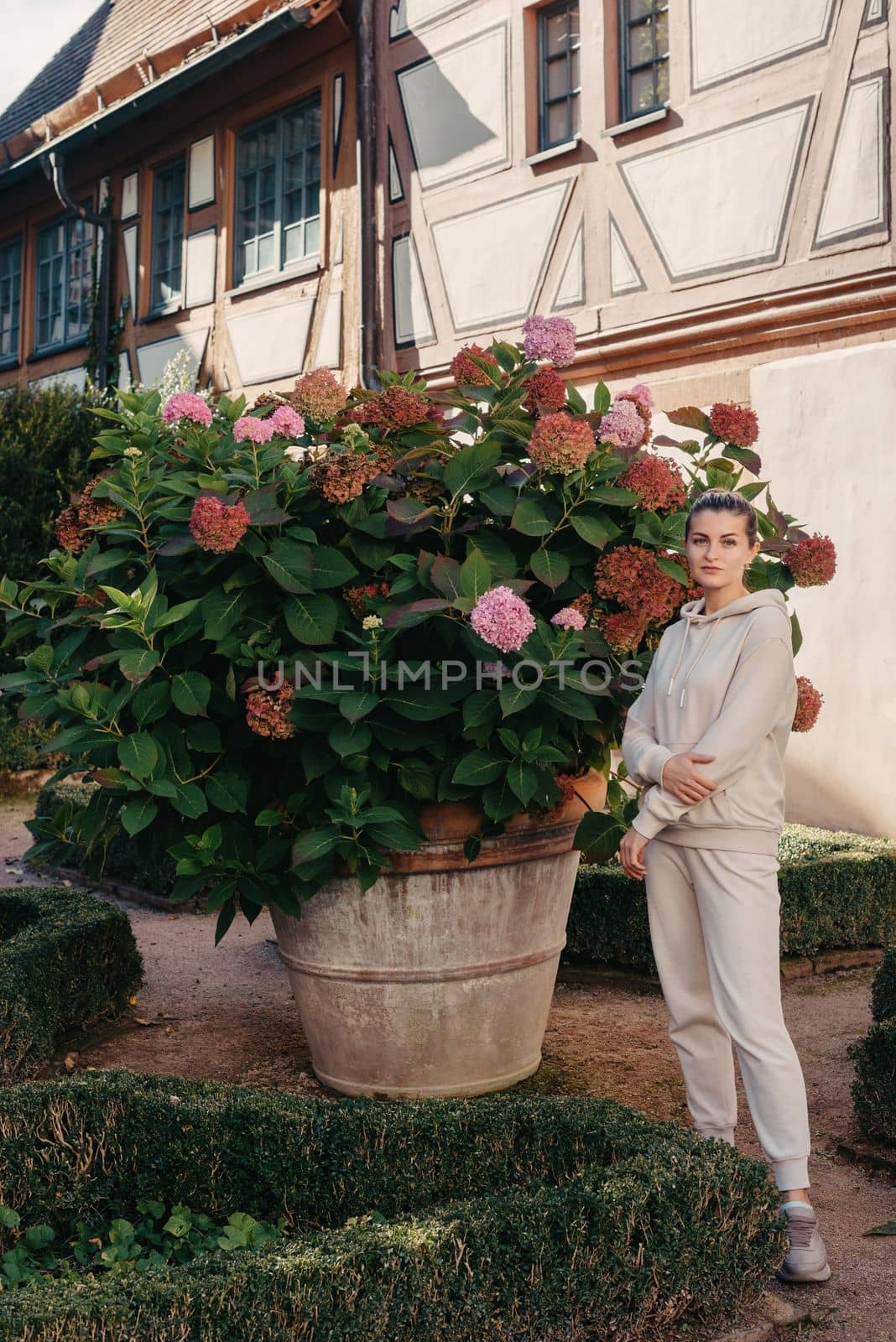 Attractive curly blonde woman walk on the city park street. Girl wear purple hoodie look happy and smiles. Woman make here me gesture standing near pink blooming bush flowers. Happy laughing girl. by Andrii_Ko