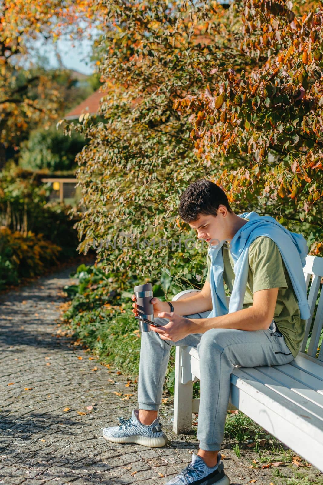 A Teenager Sits On A Bench In The Autumn Park Drinks Coffee From A Thermo Mug And Looks Into A Phone. Portrait Of Handsome Cheerful Guy Sitting On Bench Fresh Air Using Device Browsing Media Smm Drinking Latte Urban Outside Outdoor by Andrii_Ko