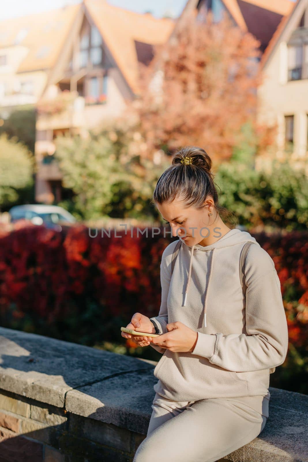 Young Fashionable Teenage Girl With Smartphone In Europian Park In Autumn Sitting At Smiling. Trendy Young Woman In Fall In Park Texting. Retouched, Vibrant Colors. Beautiful Blonde Teenage Girl Wearing Casual Modern Autumn Outfit Sitting In Park In Autumn. Retouched, Vibrant Colors, Brownish Tones. by Andrii_Ko