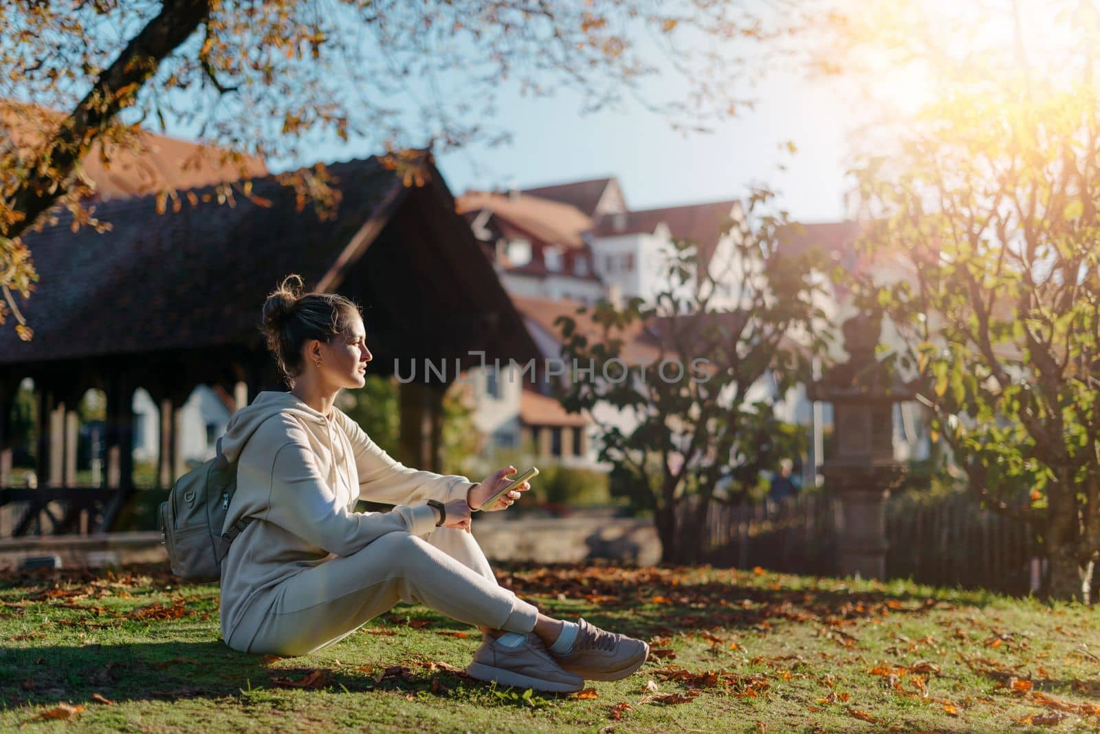 Young fashionable teenage girl with smartphone in park in autumn sitting at smiling. Trendy young woman in fall in park texting. Retouched, vibrant colors. Beautiful blonde teenage girl wearing casual modern autumn outfit sitting in park in autumn. Retouched, vibrant colors, brownish tones.