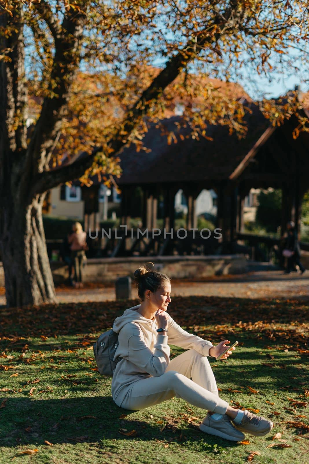 Young fashionable teenage girl with smartphone in park in autumn sitting at smiling. Trendy young woman in fall in park texting. Retouched, vibrant colors. Beautiful blonde teenage girl wearing casual modern autumn outfit sitting in park in autumn. Retouched, vibrant colors, brownish tones.