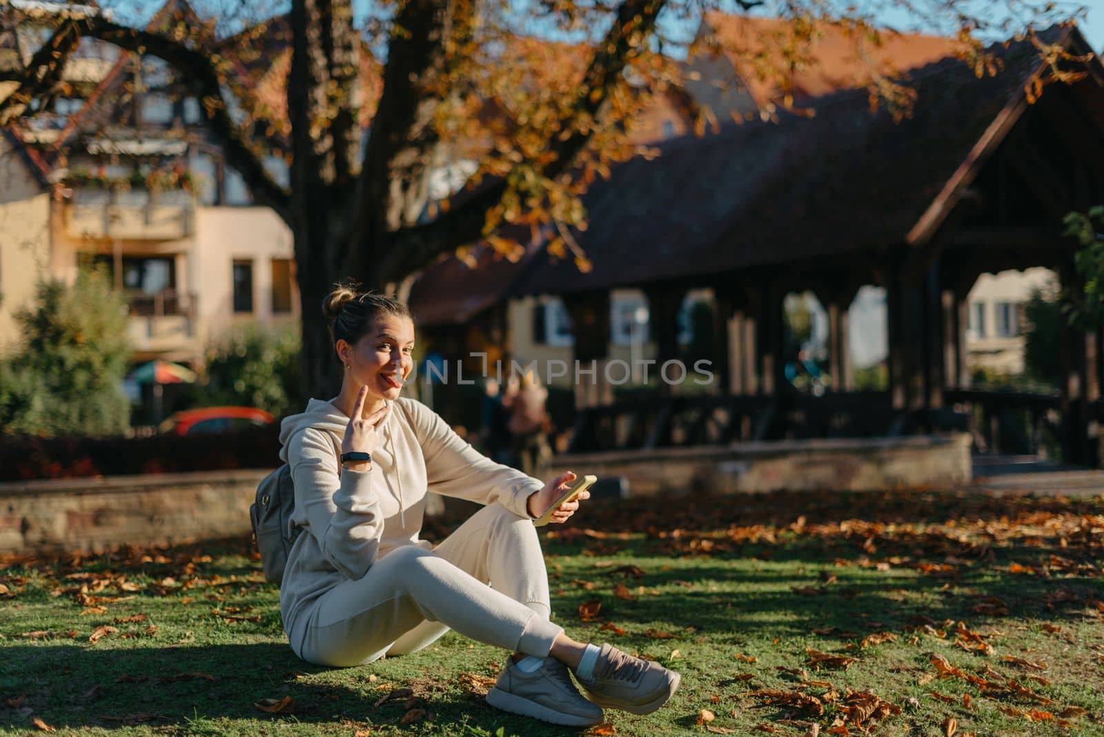 Young fashionable teenage girl with smartphone in park in autumn sitting at smiling. Trendy young woman in fall in park texting. Retouched, vibrant colors. Beautiful blonde teenage girl wearing casual modern autumn outfit sitting in park in autumn. Retouched, vibrant colors, brownish tones.