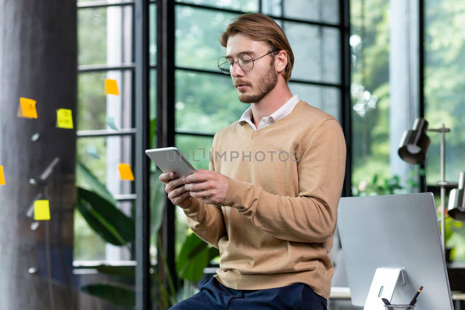 Young successful thinking programmer inside loft style office uses tablet computer, young blond man in casual clothes reads news online, serious businessman browses internet pages.