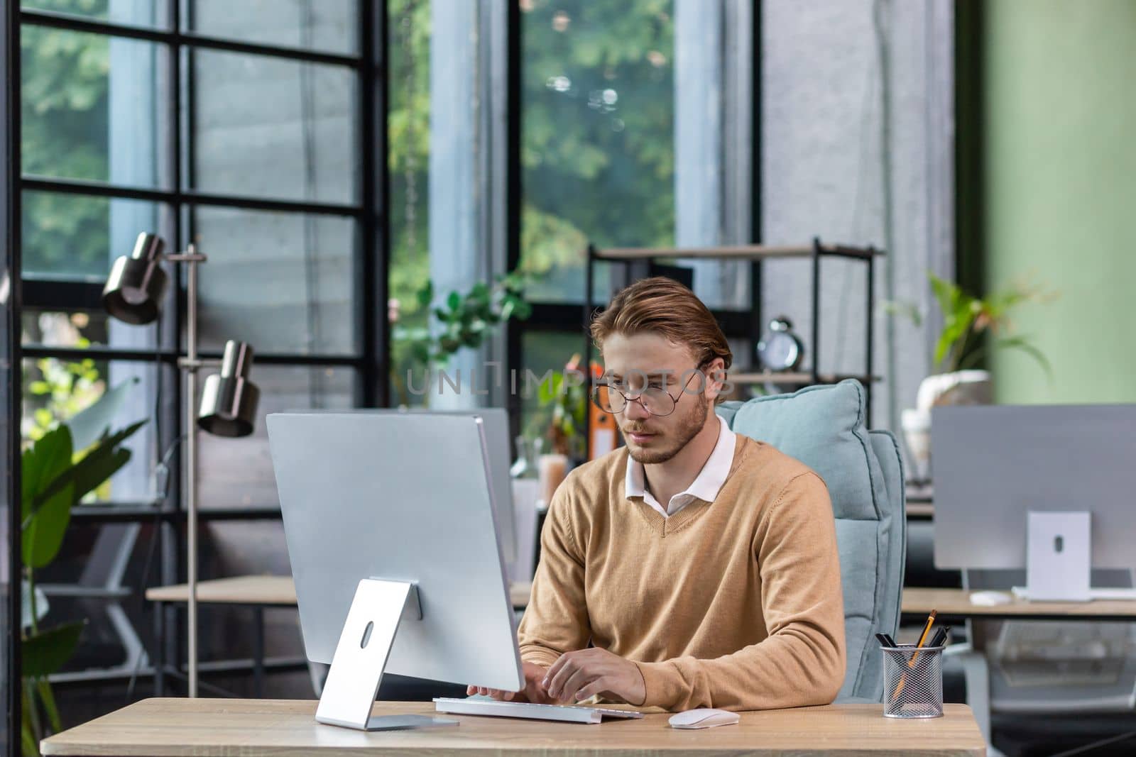 Serious and scorn-centered man in the office at work with a computer, man typing thoughtfully on keyboards, businessman young blond in sweater indoors loft, with green colors, programmer at work by voronaman