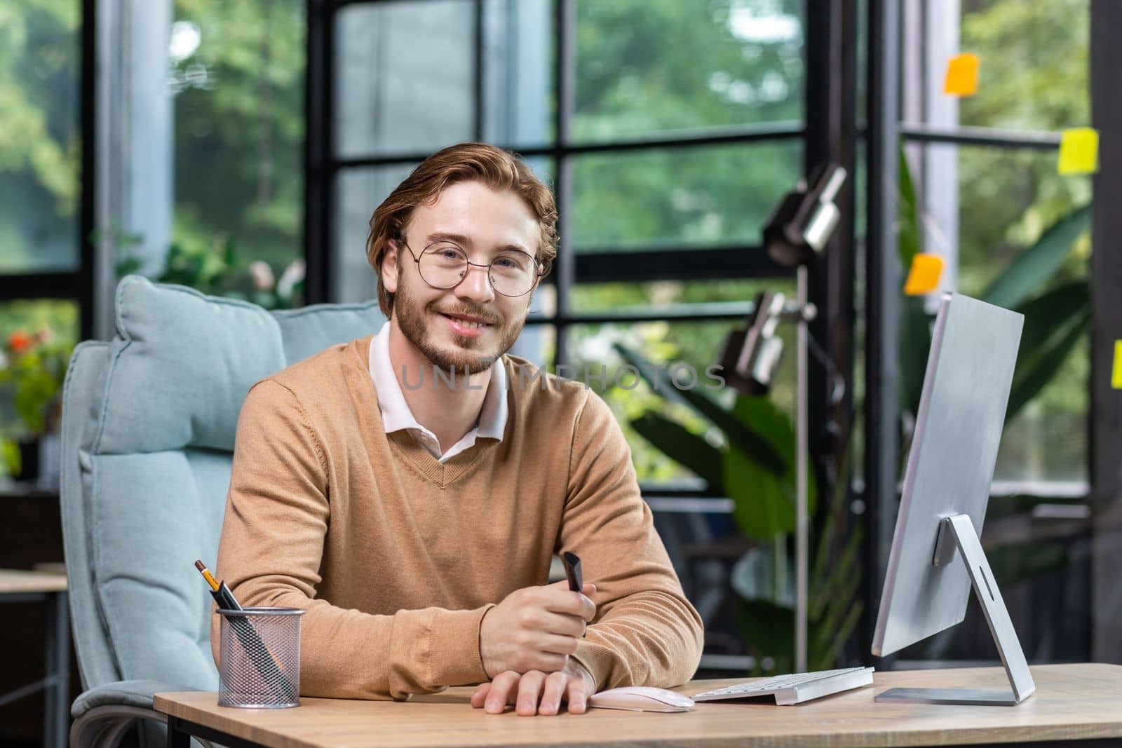 Portrait of successful smiling programmer inside modern green loft office, blond man smiling and looking at camera, businessman in sweater and casual shirt close up working with computer.