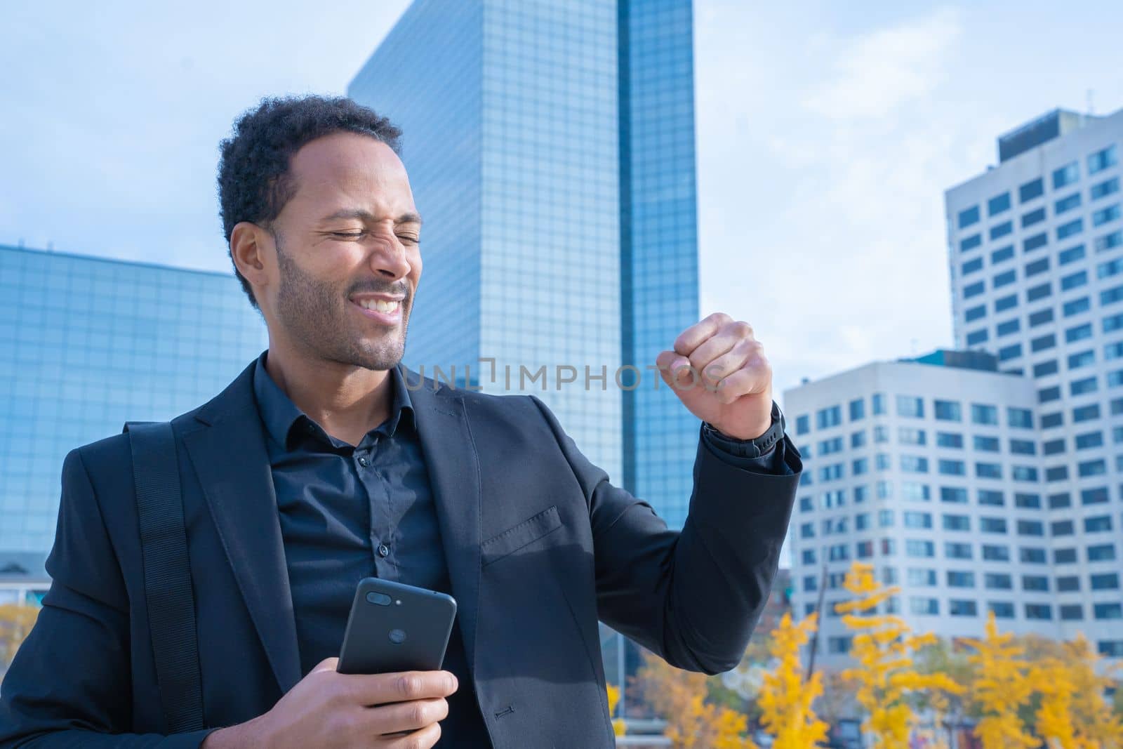 Cheerful businessman in suit raised arms smiling happy celebrates success with financial buildings in background. Happy real people. High quality photo