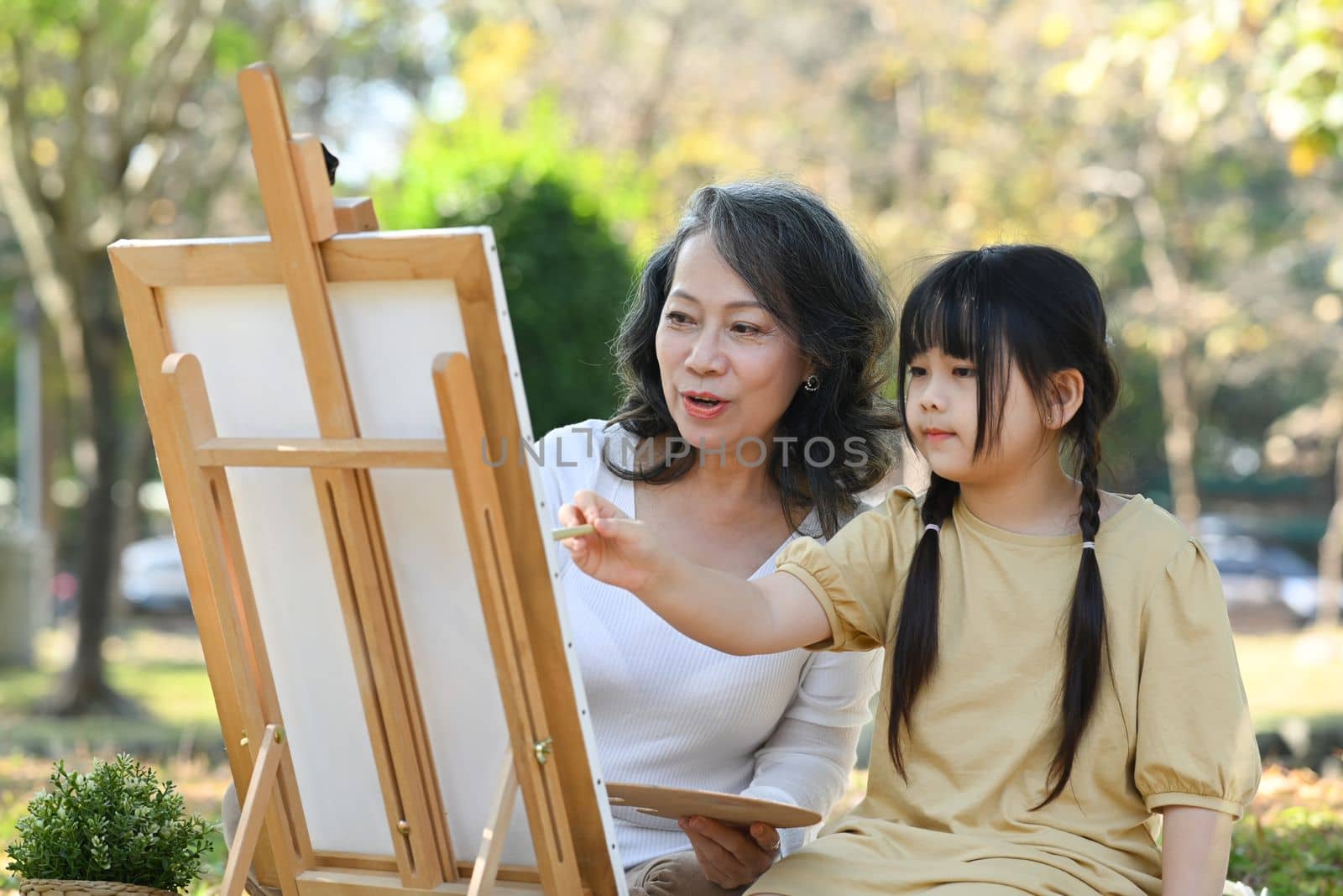 Lovely little girl and grandmother drawing in summer park, spending leisure weekend at outdoor together by prathanchorruangsak