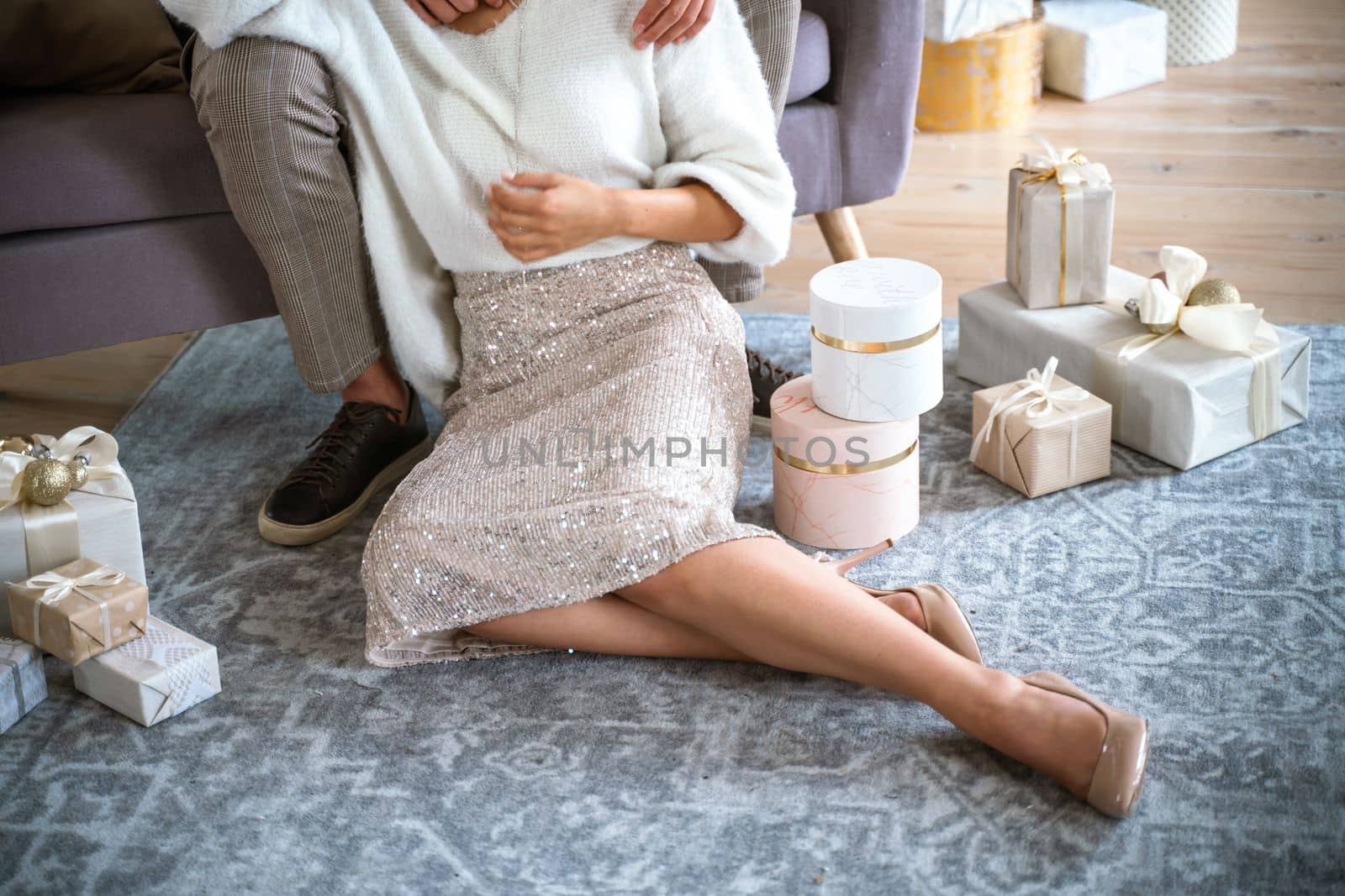 A romantic couple, a man sitting on the sofa and a woman on the carpet next to surrounded by gifts on a gray carpet. She is wearing a light skirt and beige high-heeled shoes