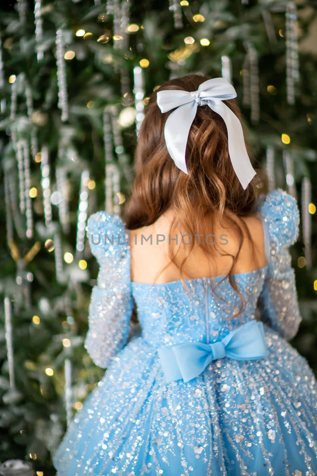 The girl decorates the Christmas tree, holding Christmas baubles in her hand. She wears a blue dress with a bow in her hair. Holiday concept