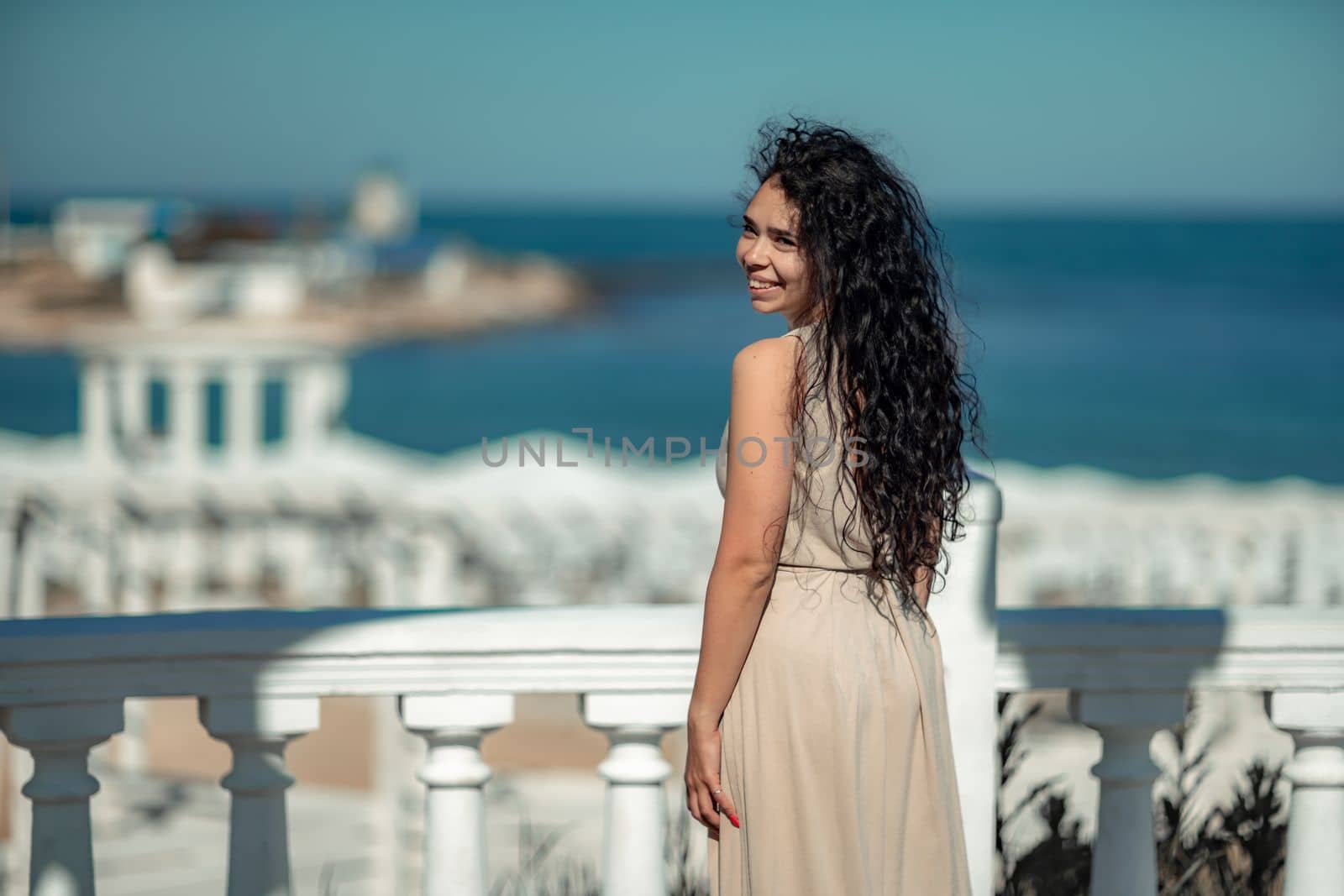 Sea woman rest. A woman with long curly hair in a beige dress stands with her back and looks at the sea and the coast from a balcony with balusters. Tourist trip to the sea
