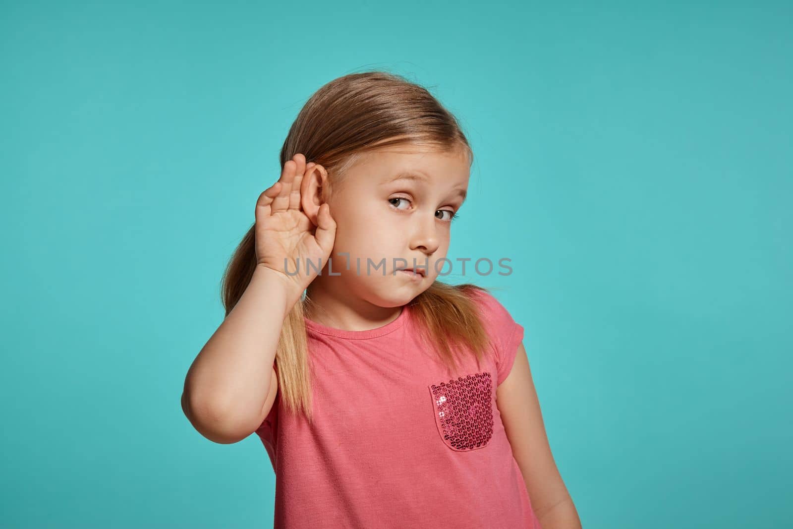 Close-up shot of a wonderful blonde little girl with two ponytails on her head, in a pink dress, acting like listening to someone and looking at the camera while posing against a blue background with copy space. Concept of a joyful childhood.