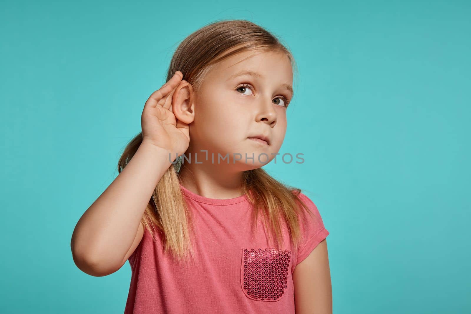 Close-up shot of a nice blonde little female with two ponytails on her head, in a pink dress, acting like listening to something while posing against a blue background with copy space. Concept of a joyful childhood.