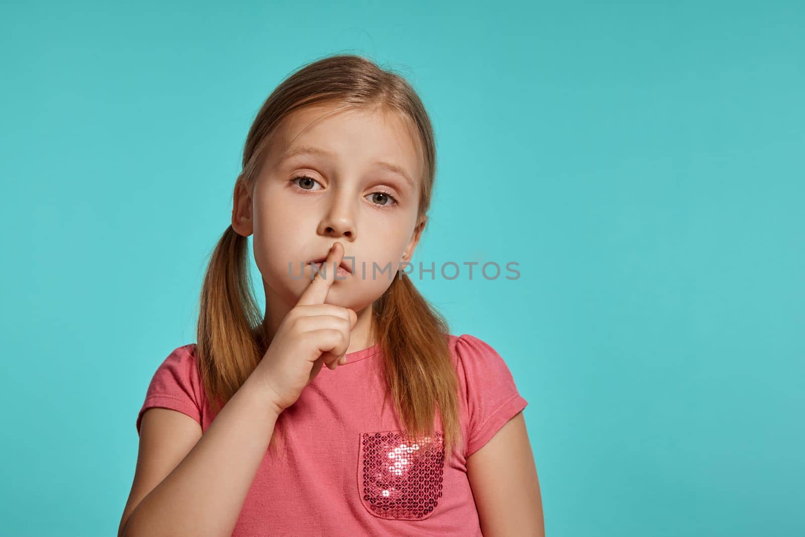 Close-up shot of a nice blonde kid with two ponytails on her head, in a pink dress, showing sign quiet and looking at the camera posing against a blue background with copy space. Concept of a joyful childhood.