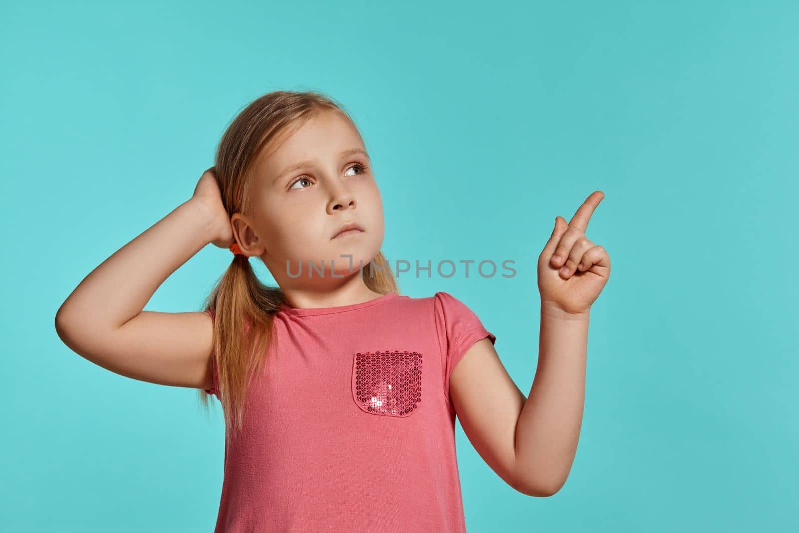 Close-up shot of a pretty blonde little female with two ponytails on her head, in a pink dress, pointing on something and looking up while posing against a blue background with copy space. Concept of a joyful childhood.