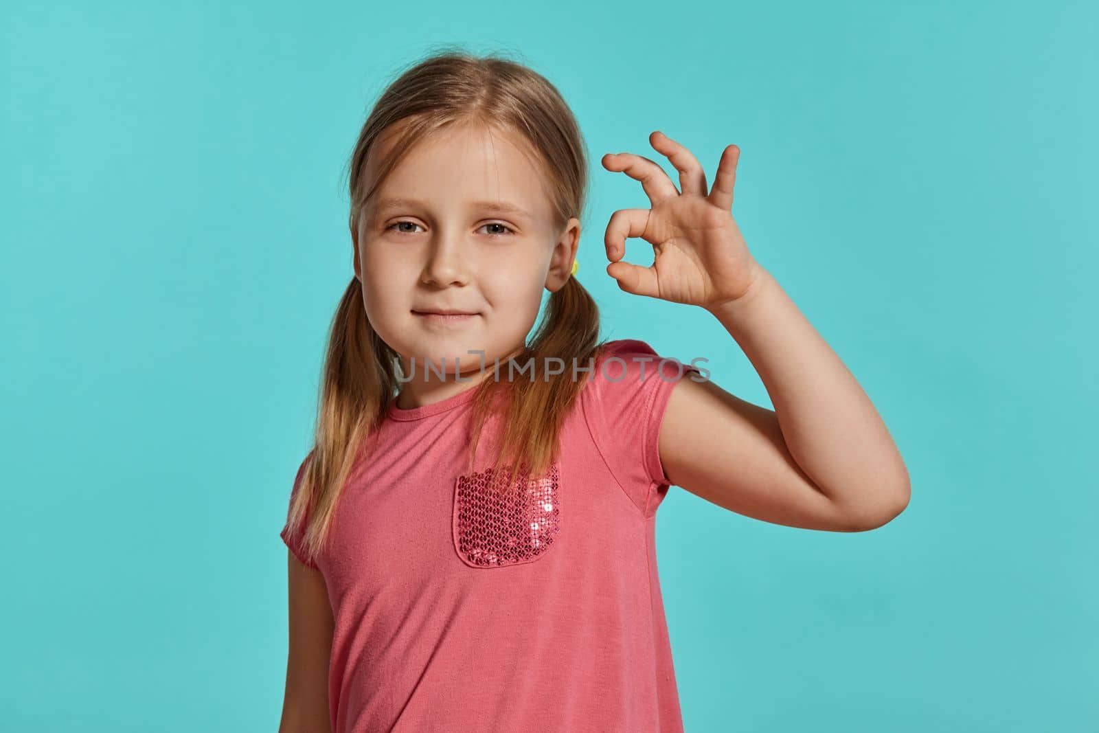 Close-up shot of a beautiful blonde little female with two ponytails on her head, in a pink dress, showing ok sign against a blue background with copy space. Concept of a joyful childhood.