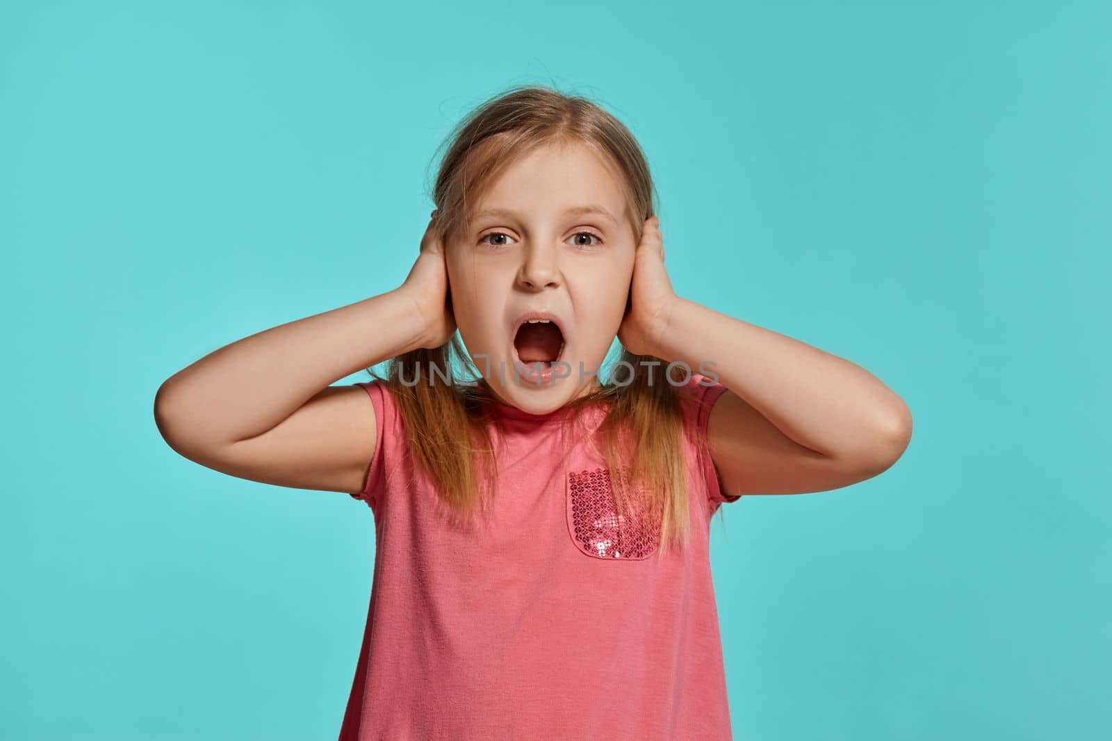 Close-up shot of beautiful blonde child with two ponytails on her head, in a pink dress, closed her ears with hands and screaming against a blue background with copy space. Concept of a joyful childhood.