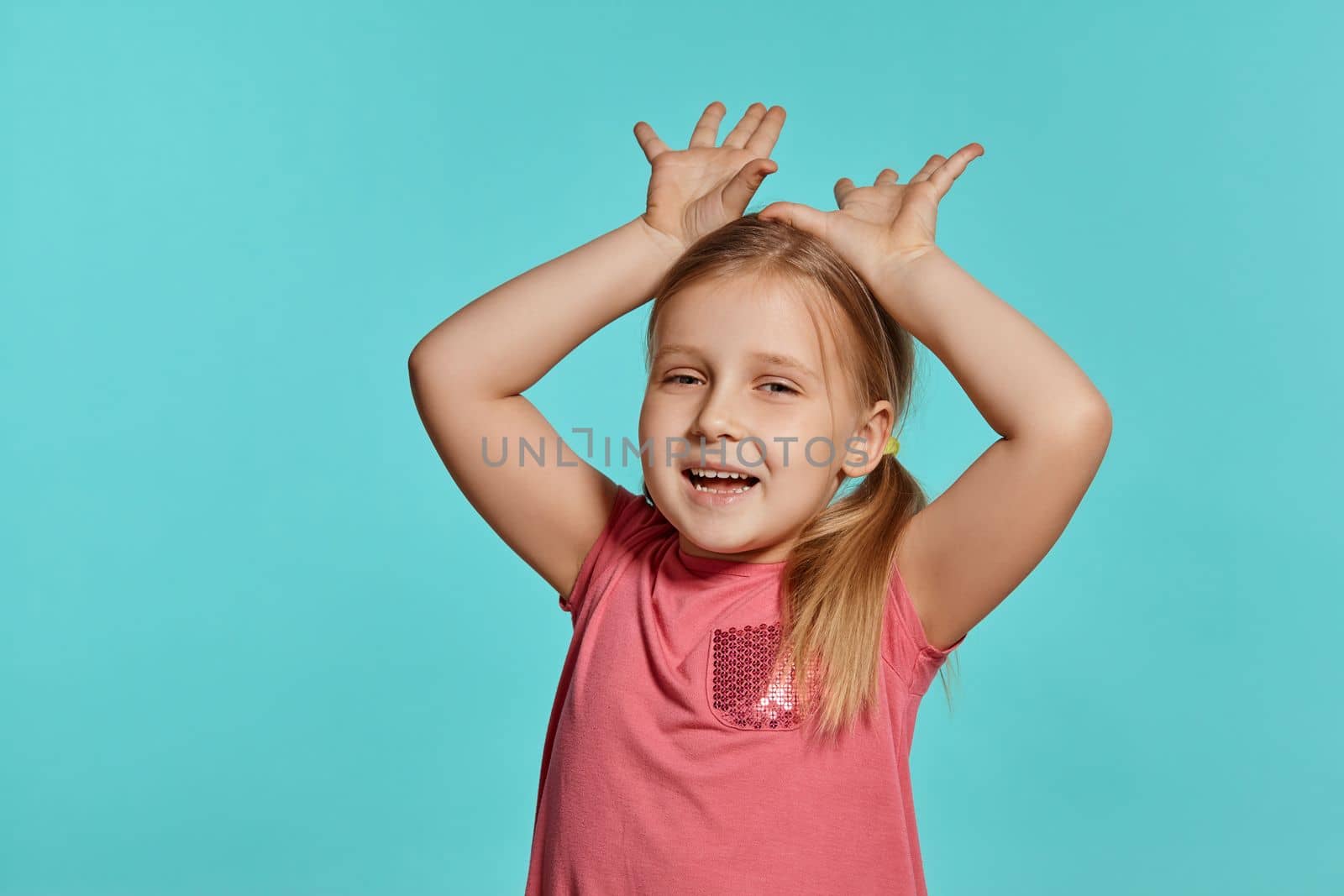 Close-up shot of a beautiful blonde little female with two ponytails on her head, in a pink dress, is making faces while posing against a blue background with copy space. Concept of a joyful childhood.