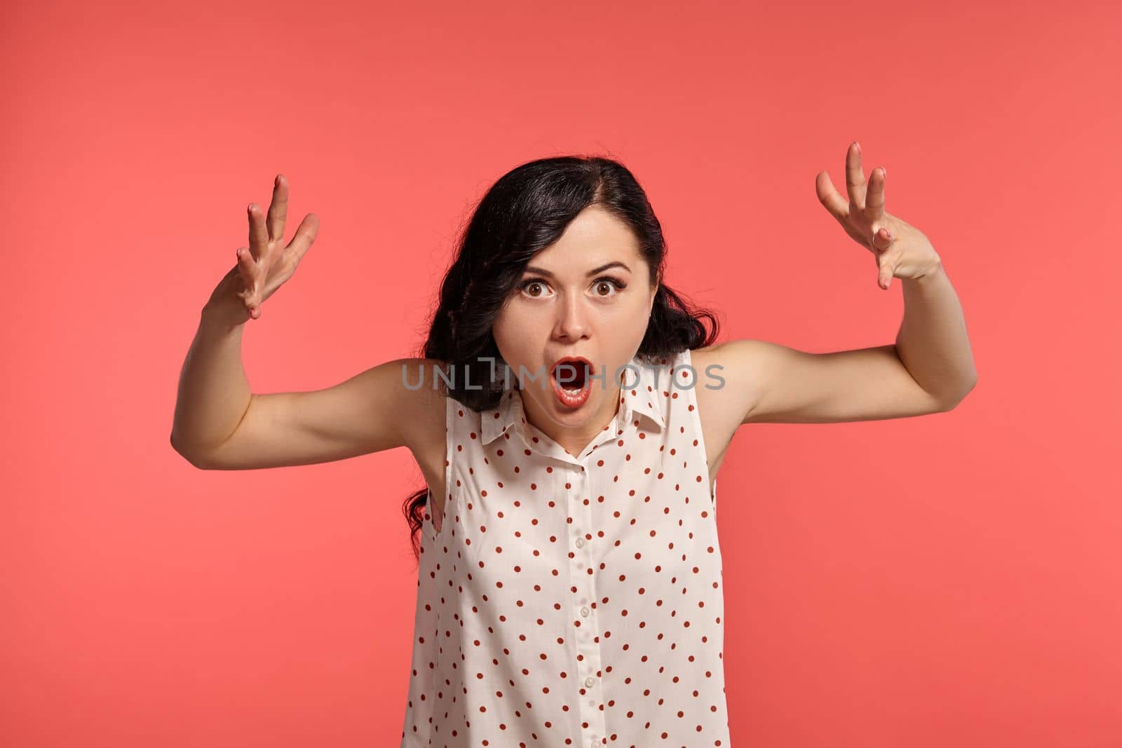 Studio shot of a pretty little woman, wearing casual white polka dot blouse. Little brunette female is acting like she is shocked, posing over a pink background. Gesticulation and sincere emotions concept.