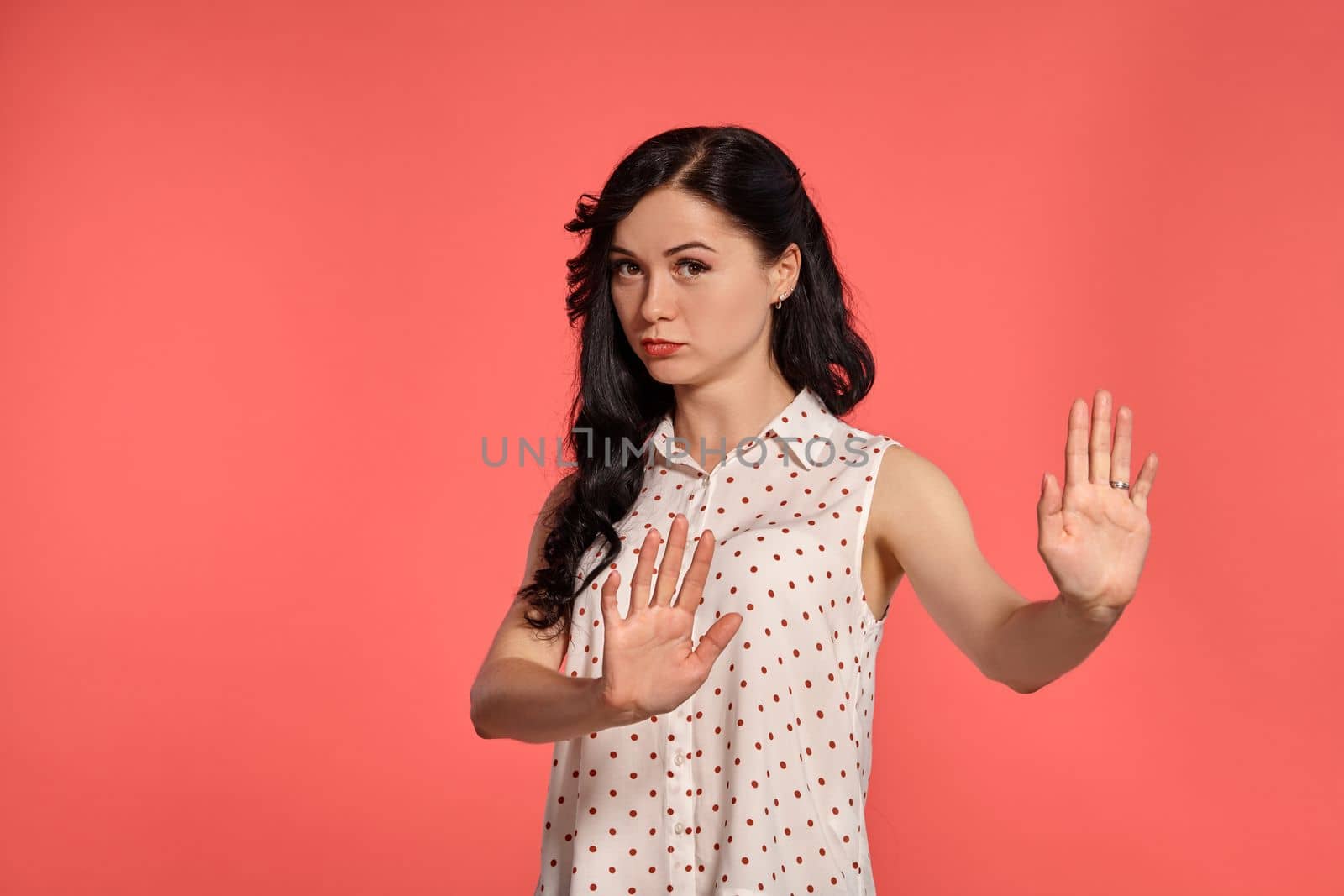 Studio shot of a pretty girl teenager, wearing casual white polka dot blouse. Little brunette female is acting like stopping somewone, posing over a pink background. Gesticulation and sincere emotions concept.