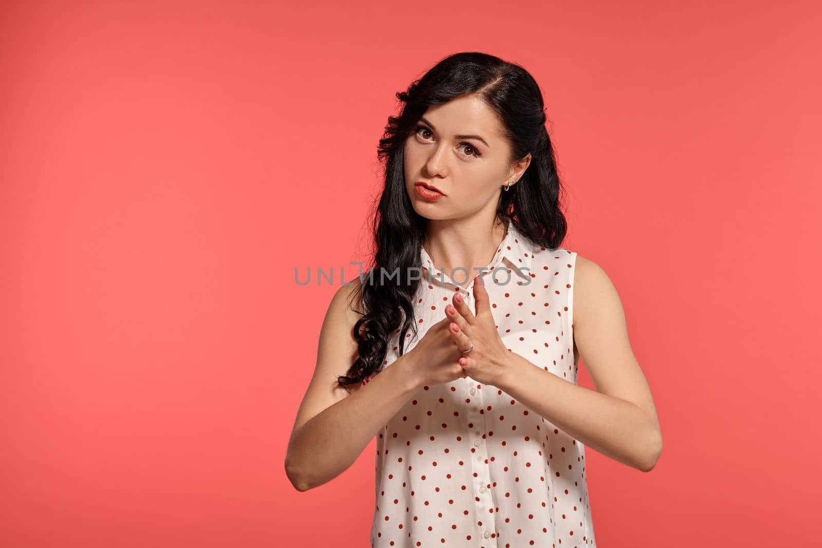 Studio shot of a cute teeny girl, wearing casual white polka dot blouse. Little brunette female is angry, looking at the camera while posing over a pink background. Gesticulation and sincere emotions concept.