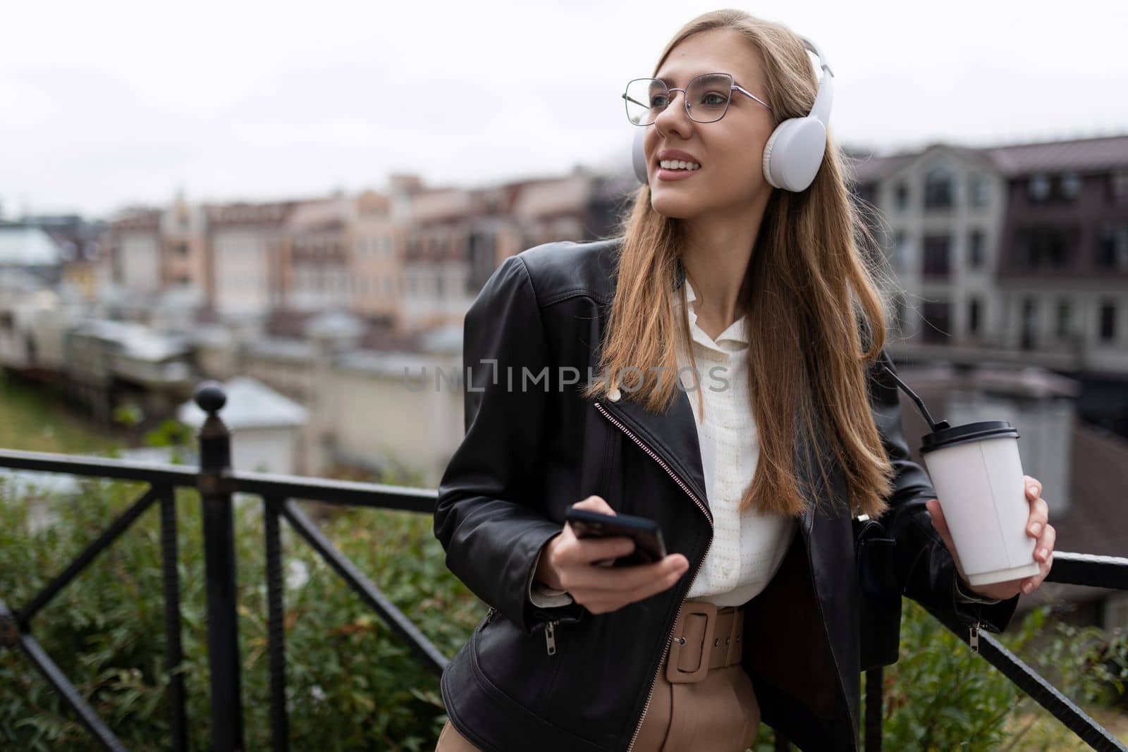 stylish young female student in headphones holding a phone and a cup of coffee in her hands outside.
