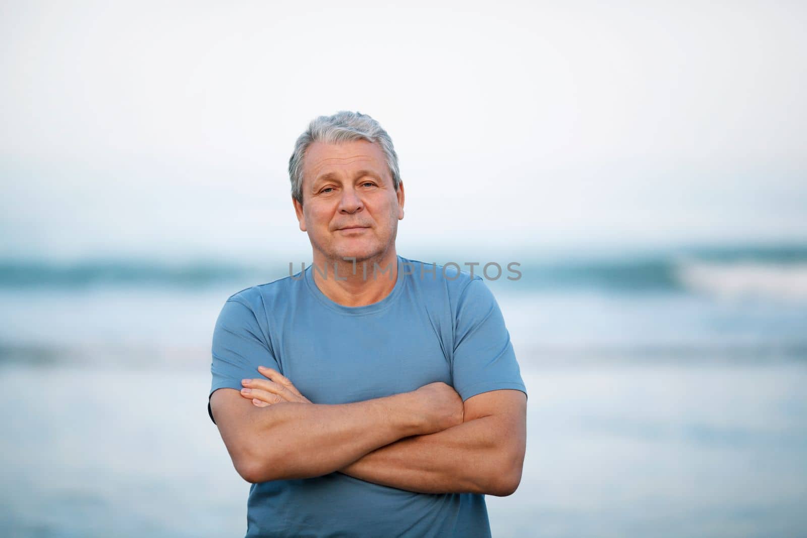 Middle shot of a senior man in blue t-shirt with arms crossed on the chest. Person looking to the camera with a light smile. View against blurry background of sea and sky
